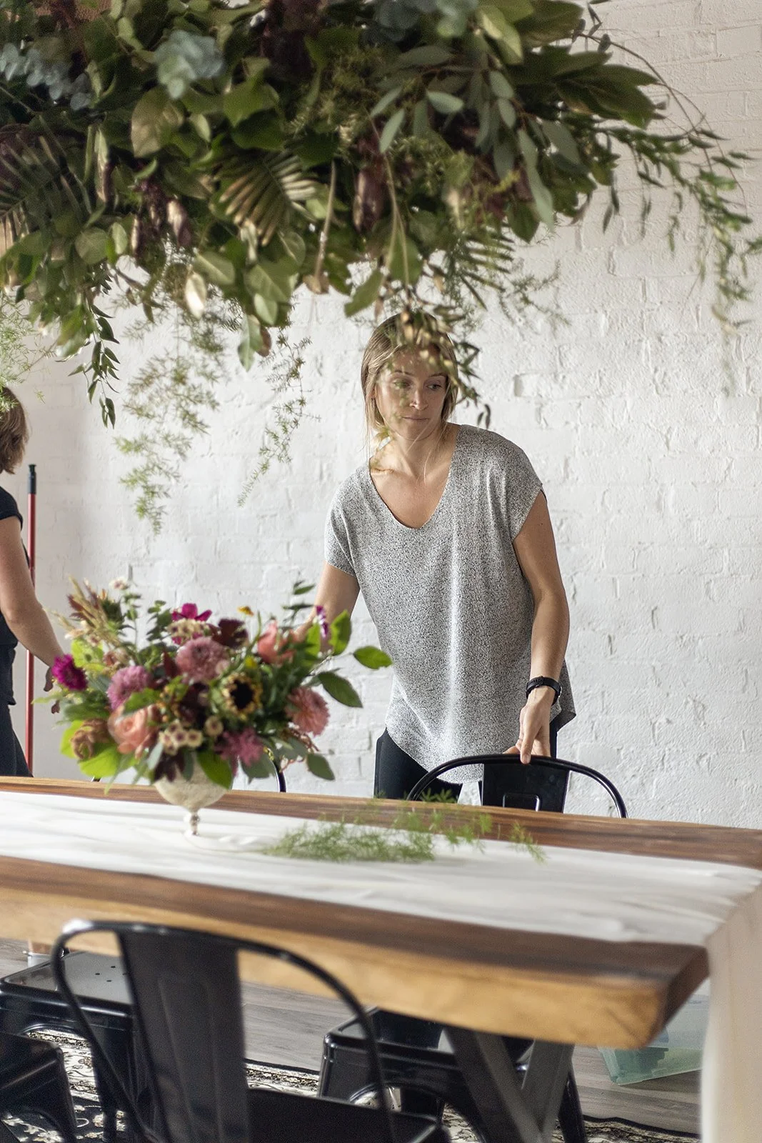 Woman arranging flowers on a wooden table decorated with a large flower arrangement, in a romantic light filled oro-medonte farm venue.