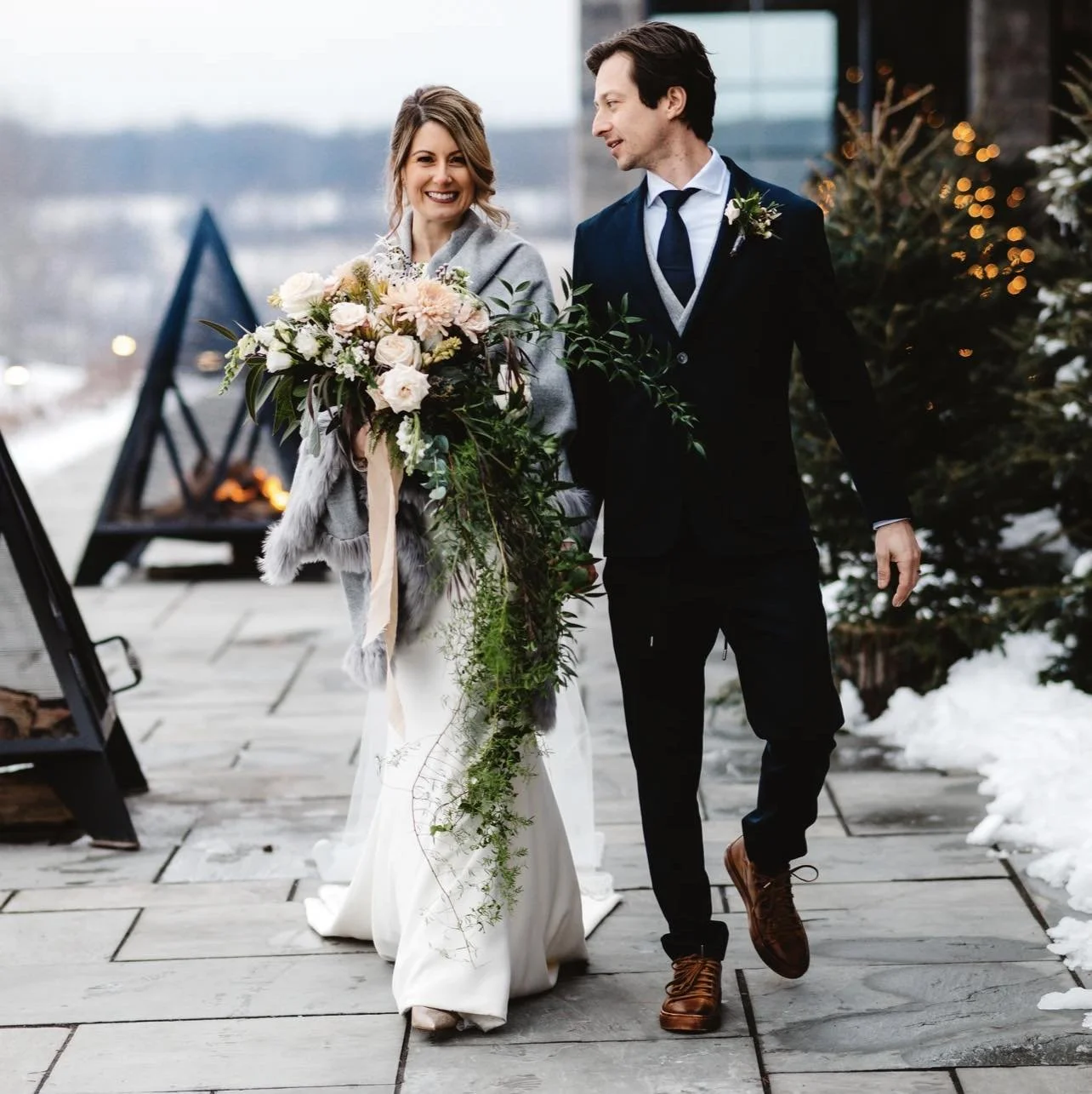 A bride and groom walking into the entrance of The KTCHN during the winter months with snow on the ground, decorated with Christmas lights, the bride holding a large bouquet of flowers.