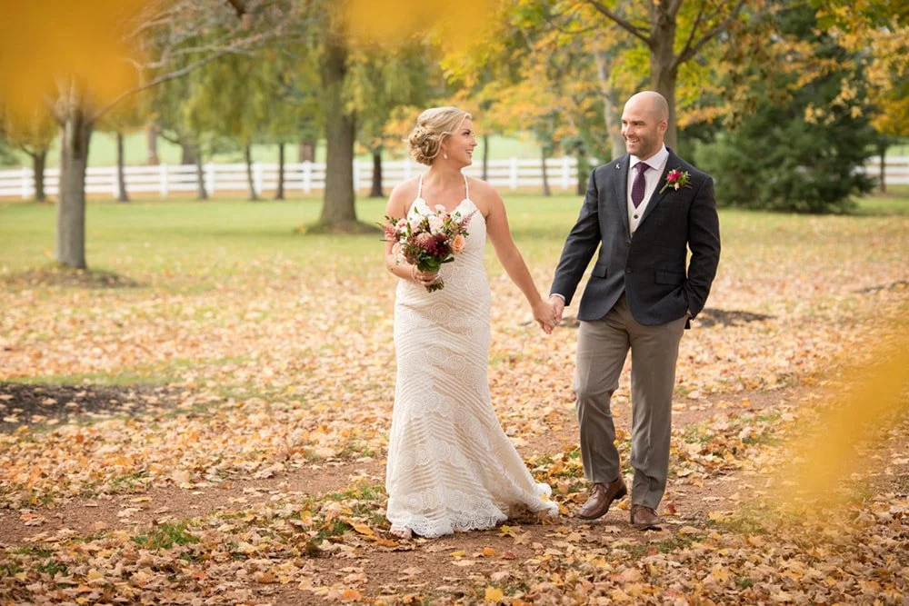 A couple in wedding attire walking hand in hand through Belcroft Estate Farm wedding venue with autumn leaves on the ground and trees with yellow and orange foliage lining the driveway.