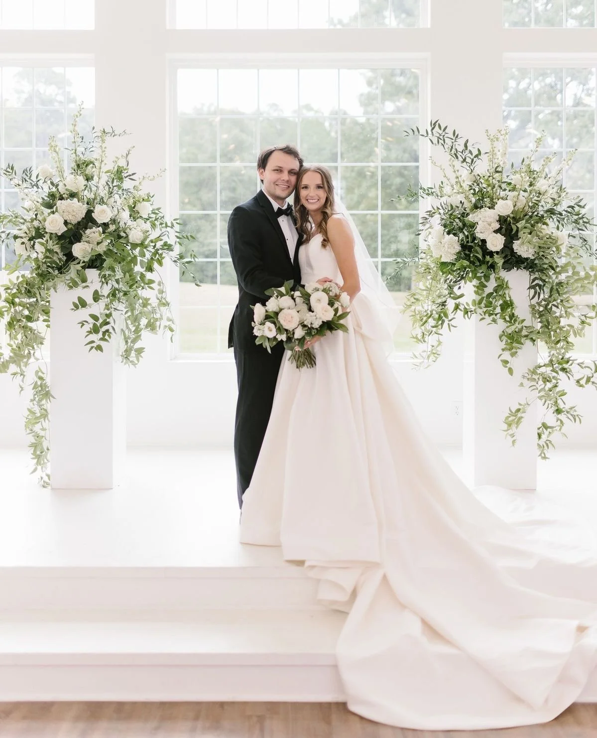 A bride and groom standing together on their wedding day, smiling, with large floral arrangements on either side, in front of a large window with a view of trees outside.