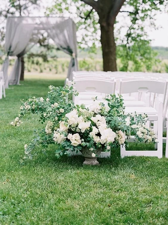 A white floral arrangement with greenery placed on the grass in front of white chairs at an outdoor event, with a tree and a white canopy in the background.