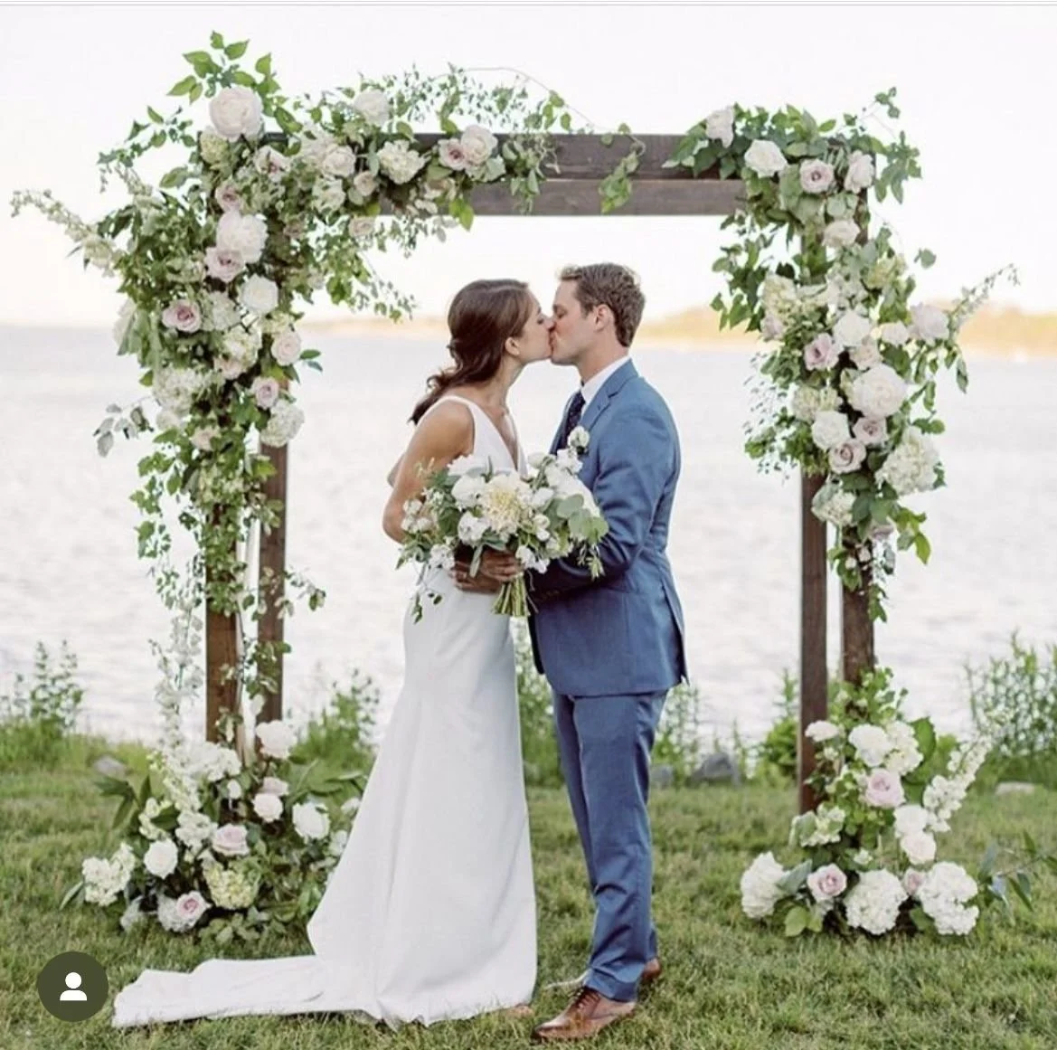 A bride and groom kiss during their outdoor wedding ceremony by a lake, framed by a floral wooden archway, with the bride holding a bouquet of white flowers and greenery, and the groom in a blue suit.