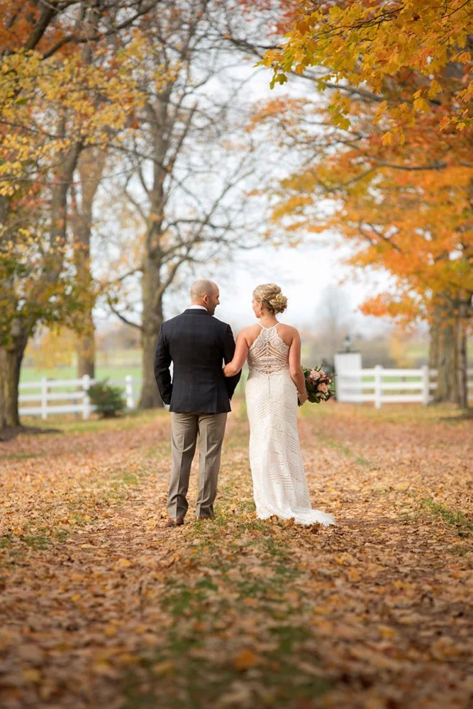 A bride and groom walking hand in hand on a leaf-covered path in an autumn park, with colorful fall trees and a white fence in the background.