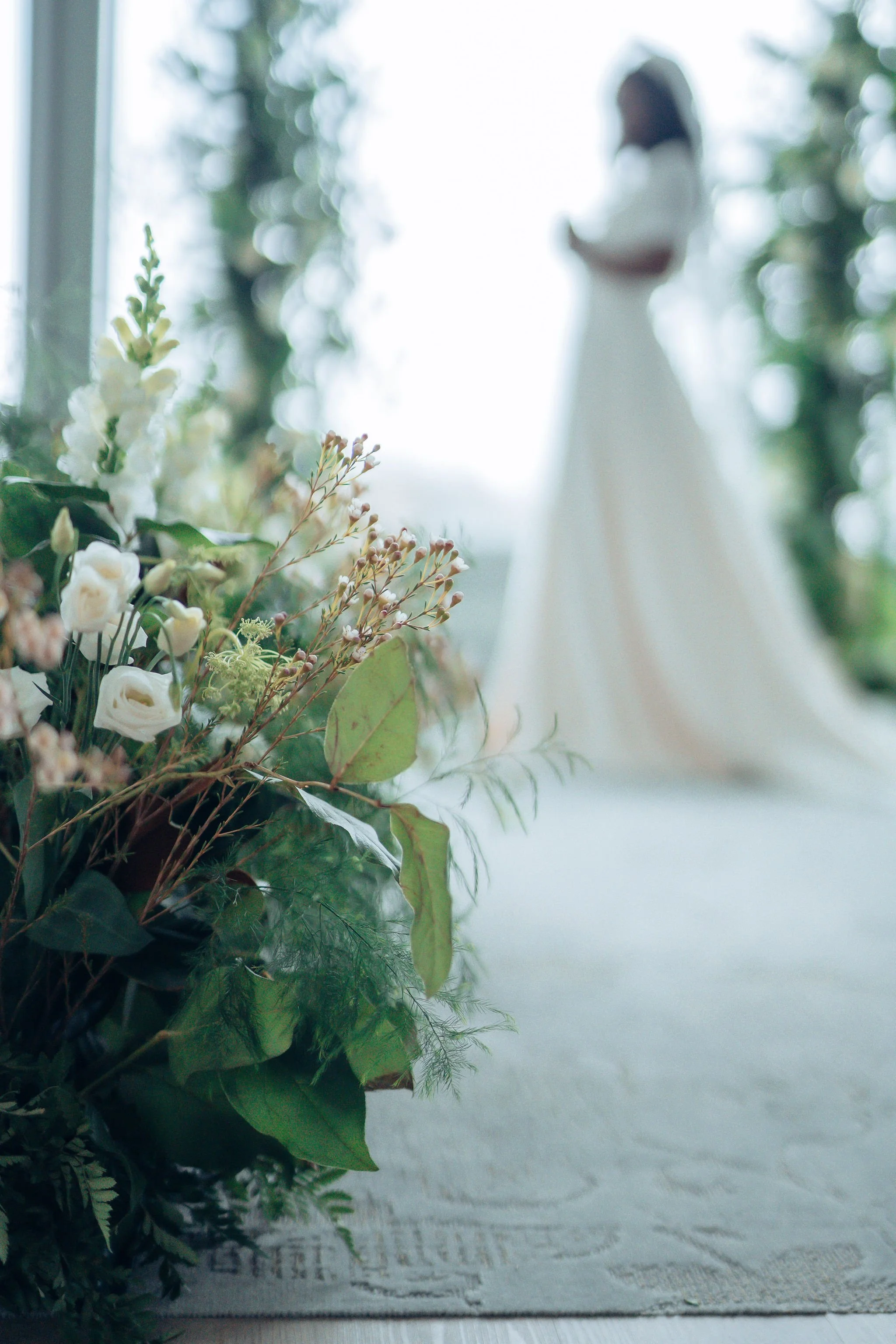 A blurred woman in a white dress stands in front of snow-covered trees, with a bouquet of white and green flowers in the foreground for her winter wedding Simcoe-Muskoka