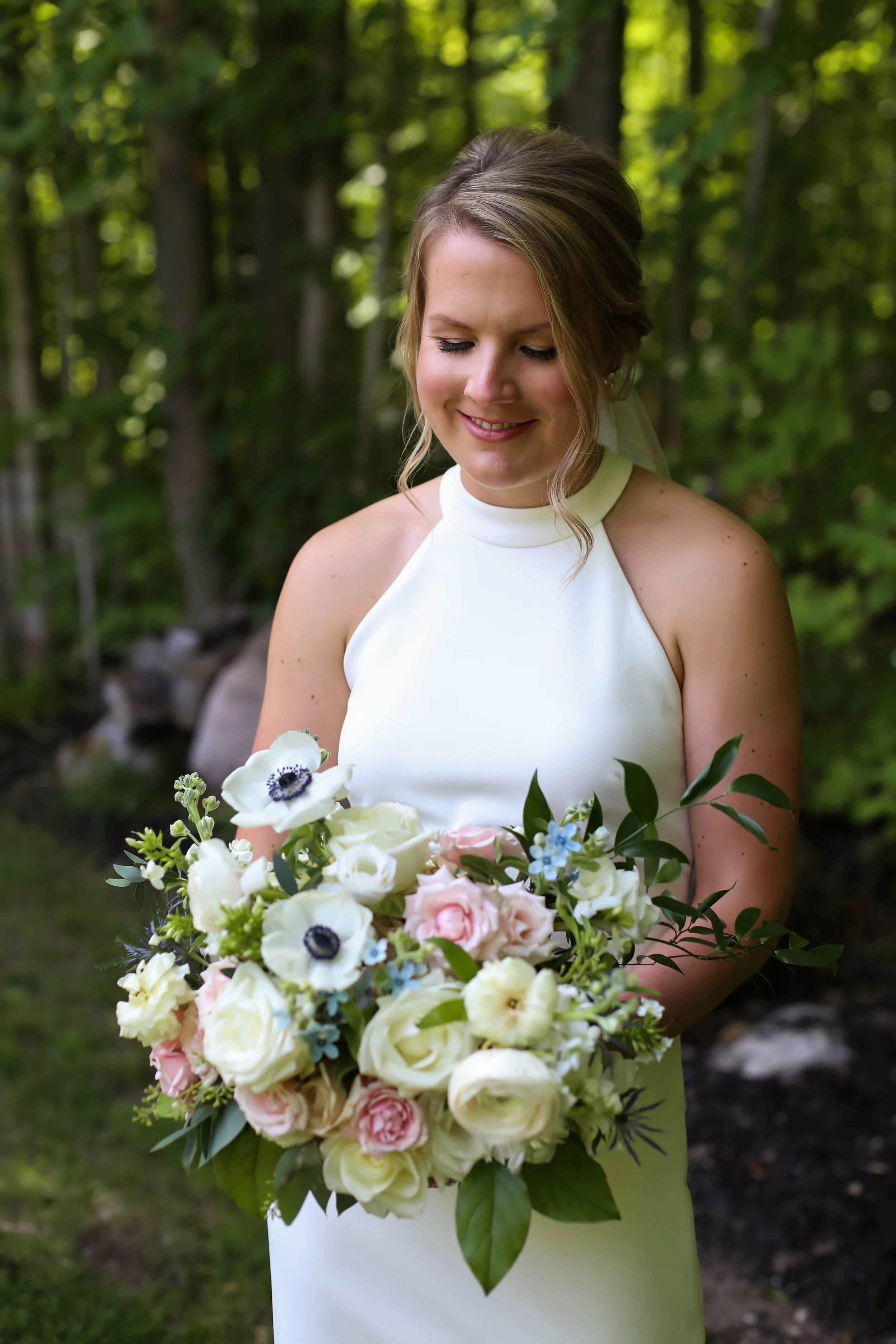 A smiling bride in a sleeveless white dress holding a large romantic bouquet of white, pink, and blue flowers, ready to get married in her outdoor ceremony location in Simcoe-Muskoka.