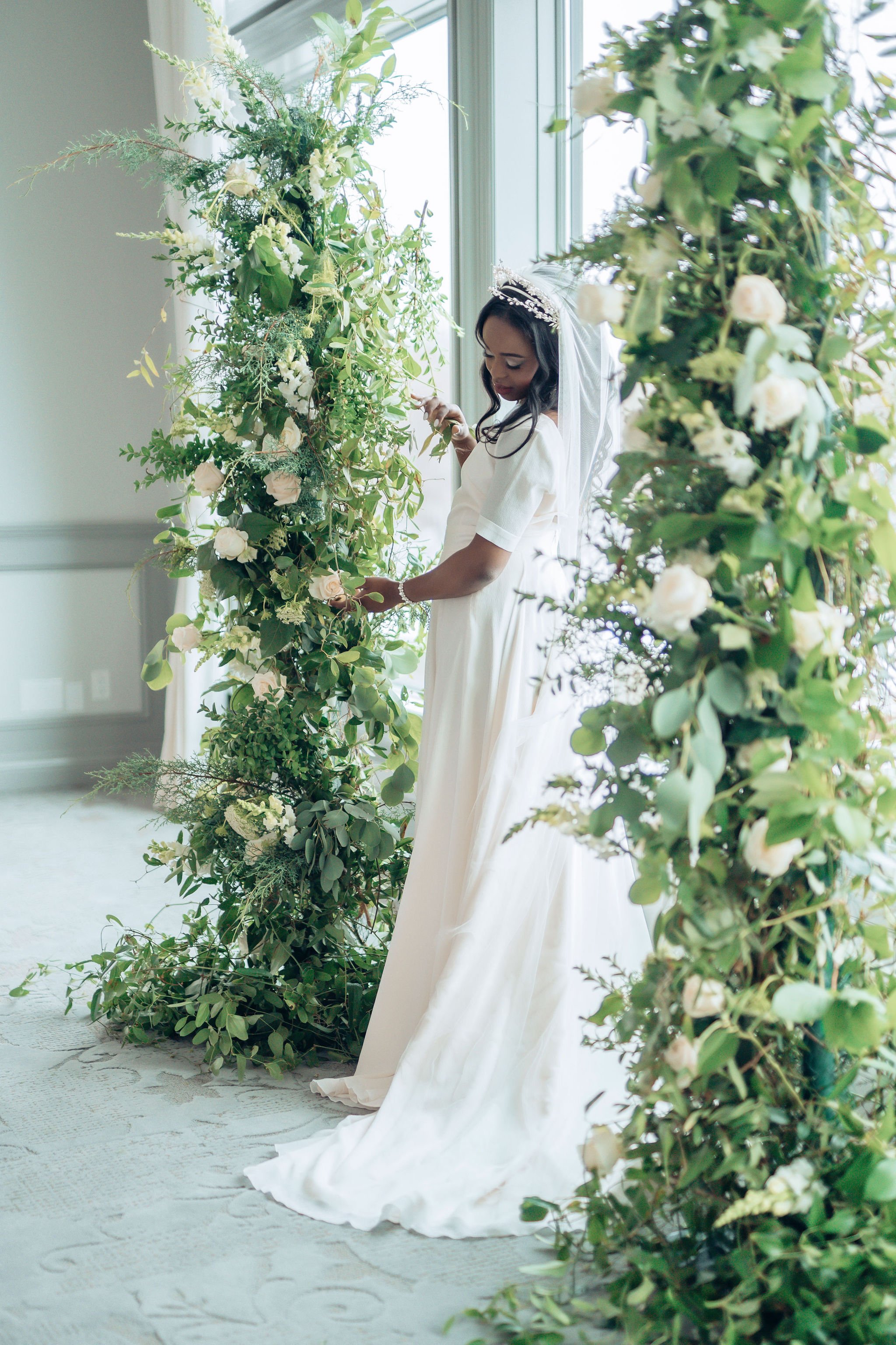 A bride in a white dress and veil stands next to a large green floral arrangement with white flowers, by a window.