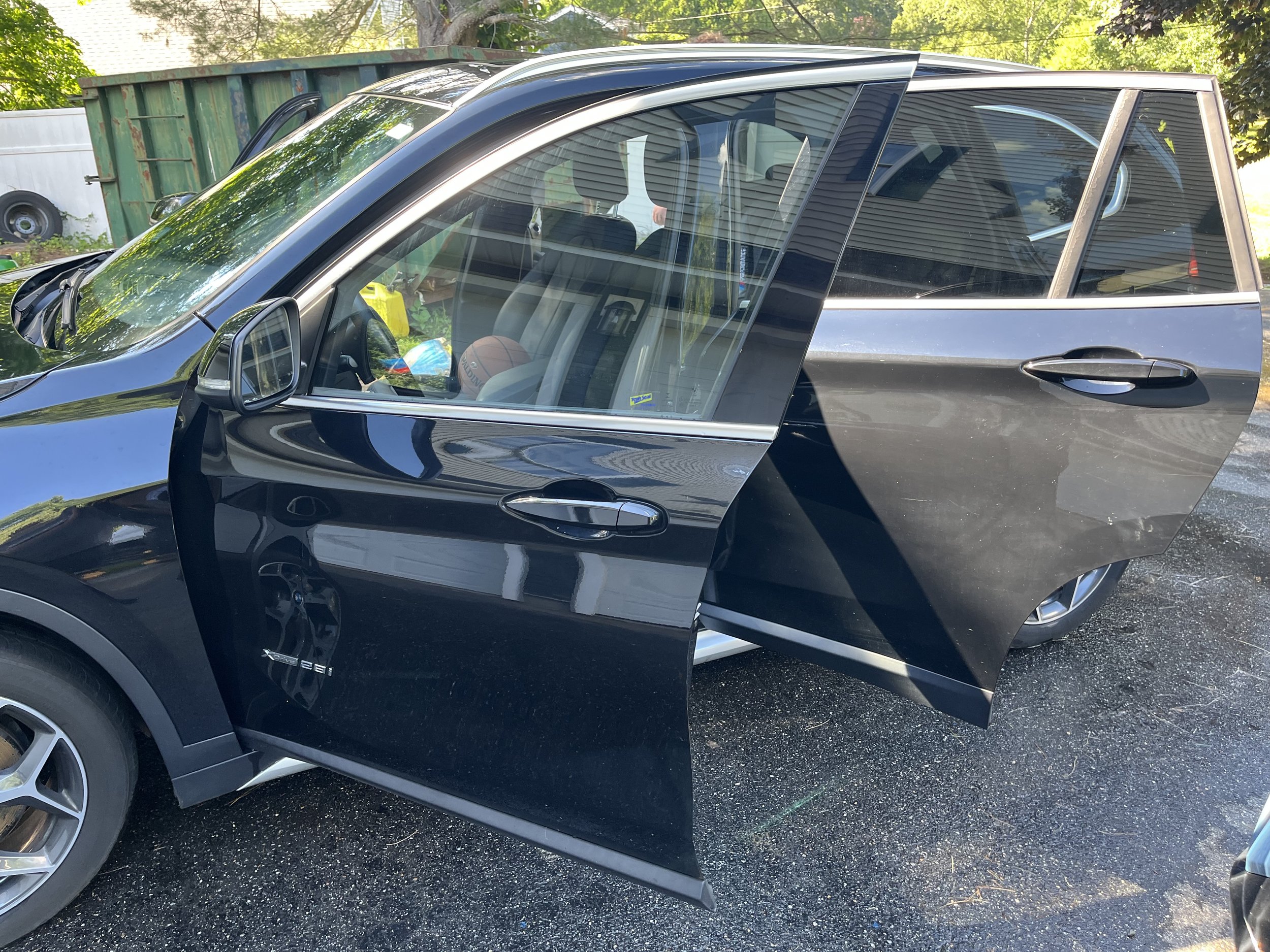 A black luxury SUV parked outdoors, with reflections of trees and a house visible on the windows. The driver's side door is slightly open, revealing the interior.