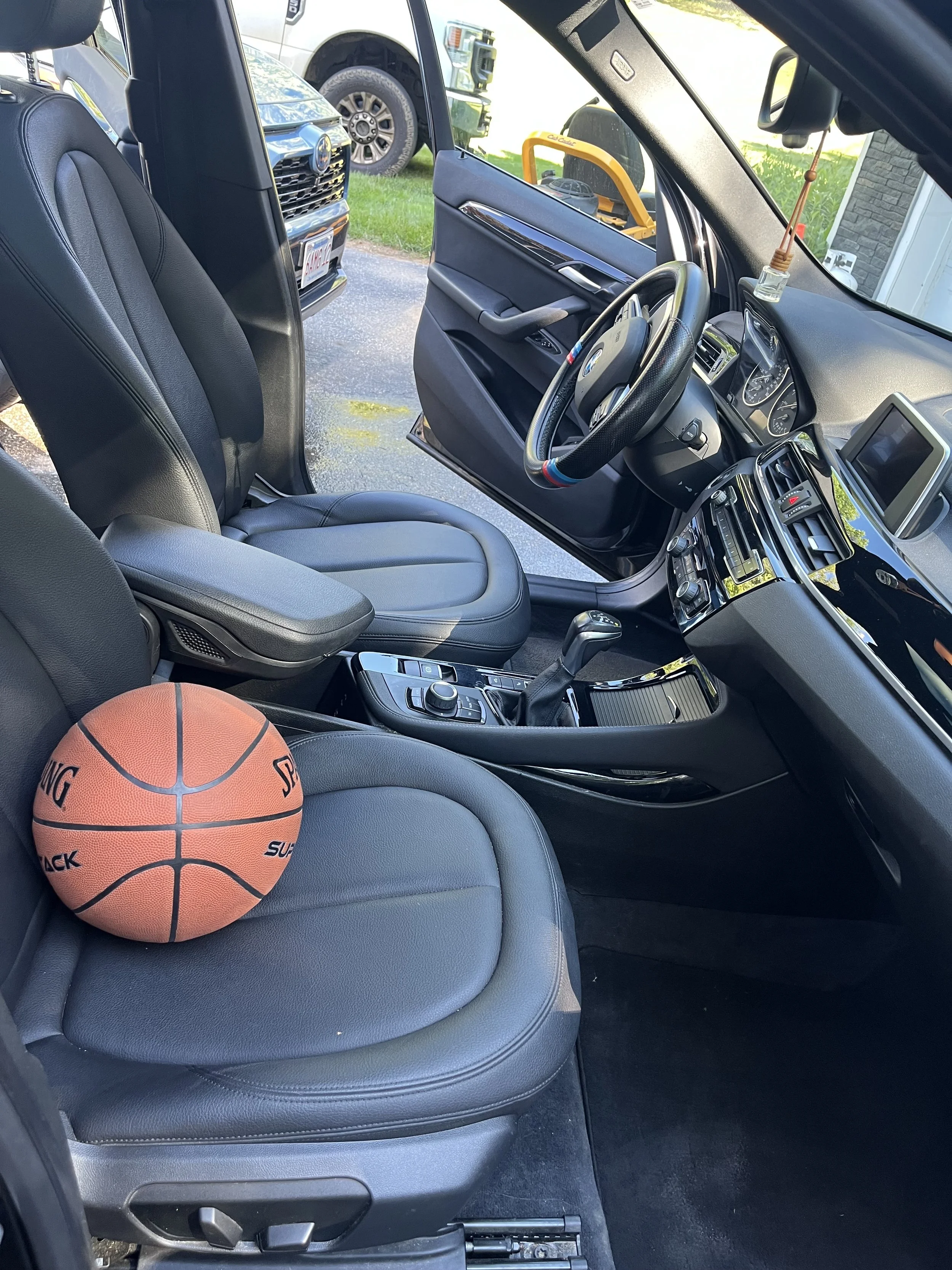The interior of a car showing the front gray seat, dashboard, steering wheel with BMW logo, and a basketball on the seat. Outside the car, there are parked vehicles and a grassy area.