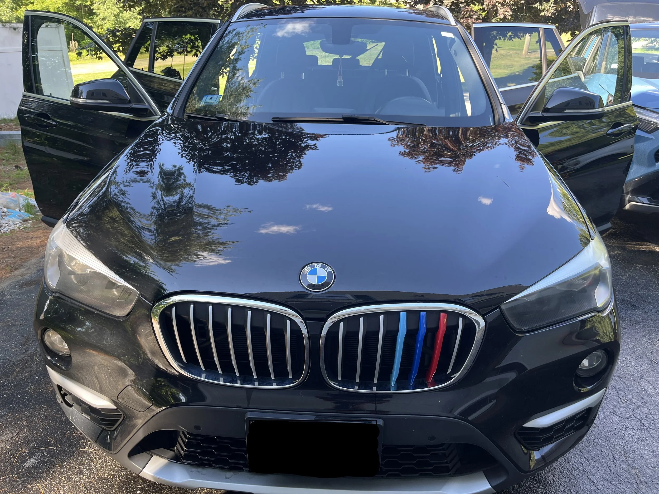 Front view of a black BMW car with open front and rear passenger doors, parked outdoors on a paved surface, with trees and greenery in the background.