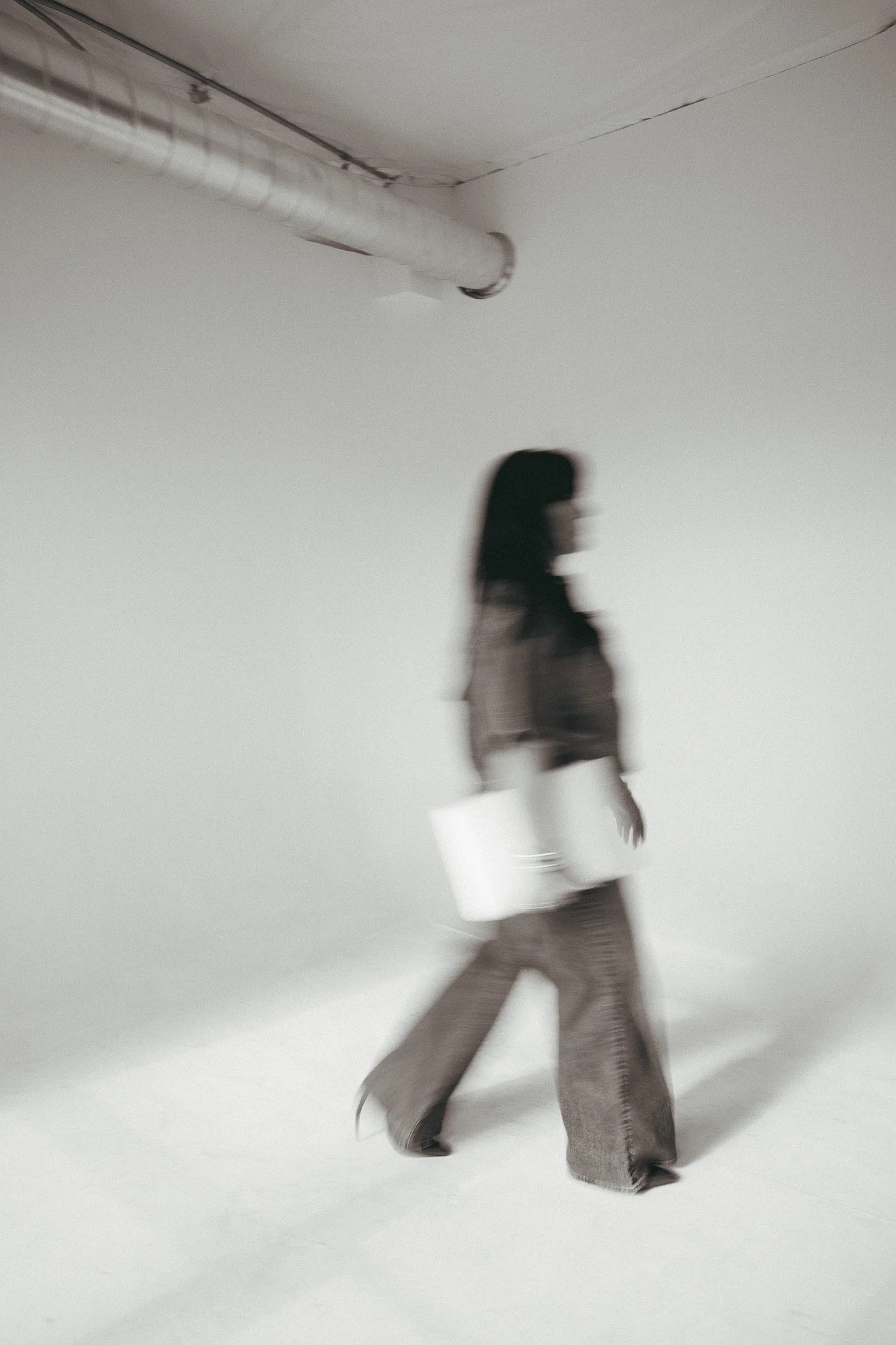 A person with dark hair wearing a beige sweater, patterned wide-leg pants, and dark shoes, walking across a plain, white background.