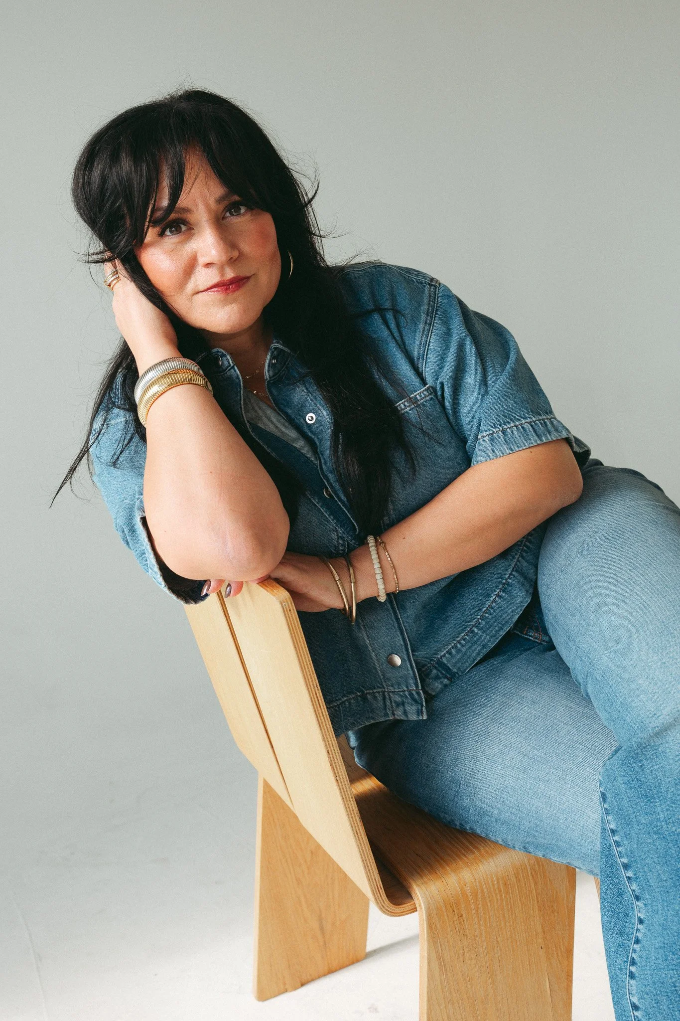Woman in a denim jacket and jeans sitting on a wooden chair with neutral gray background
