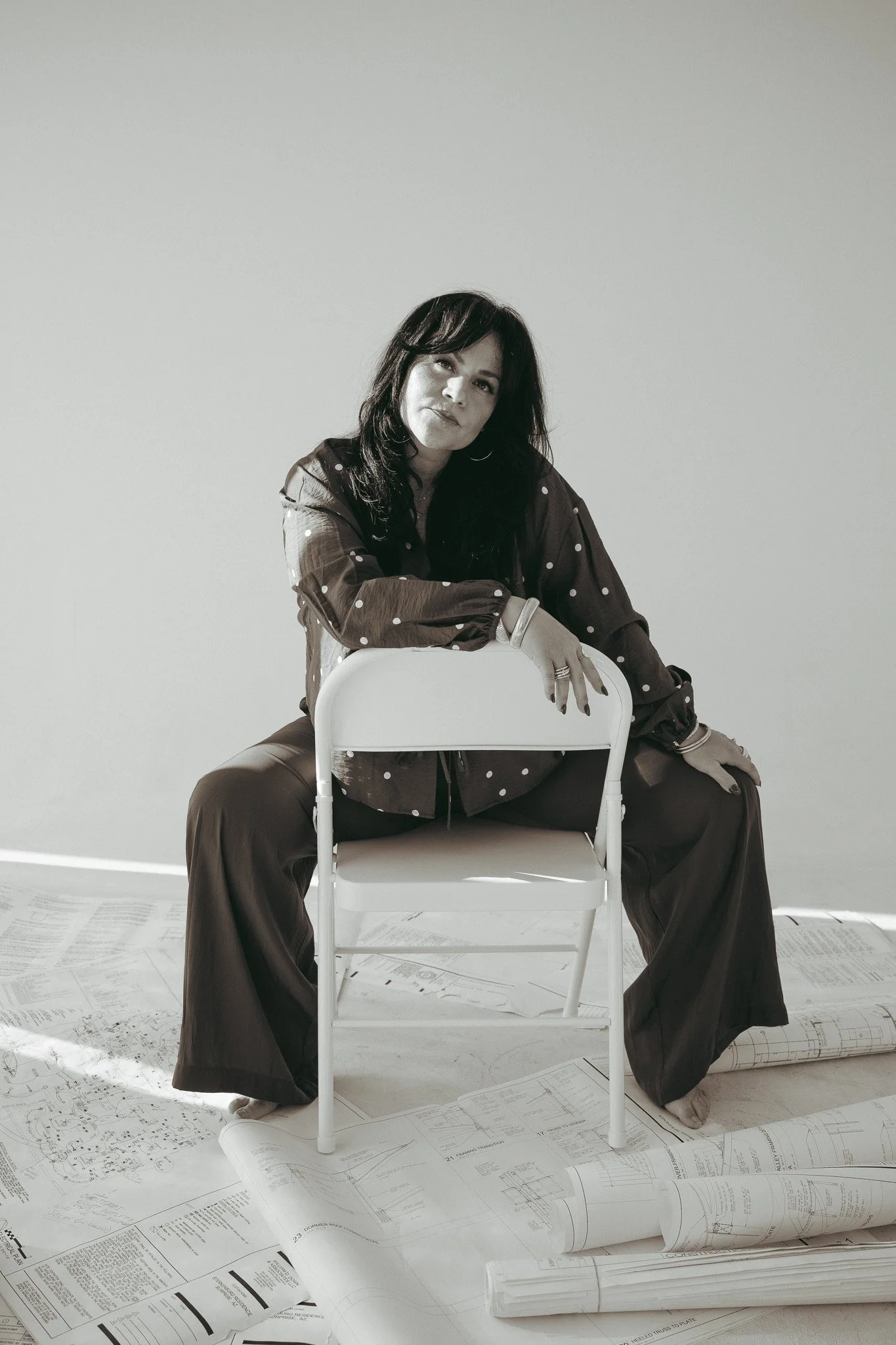 A woman with dark hair, wearing a polka dot shirt, is sitting on the floor behind a white chair, surrounded by architectural blueprints and rolled whiteprints on the floor, in a minimal room with plain light-colored walls.