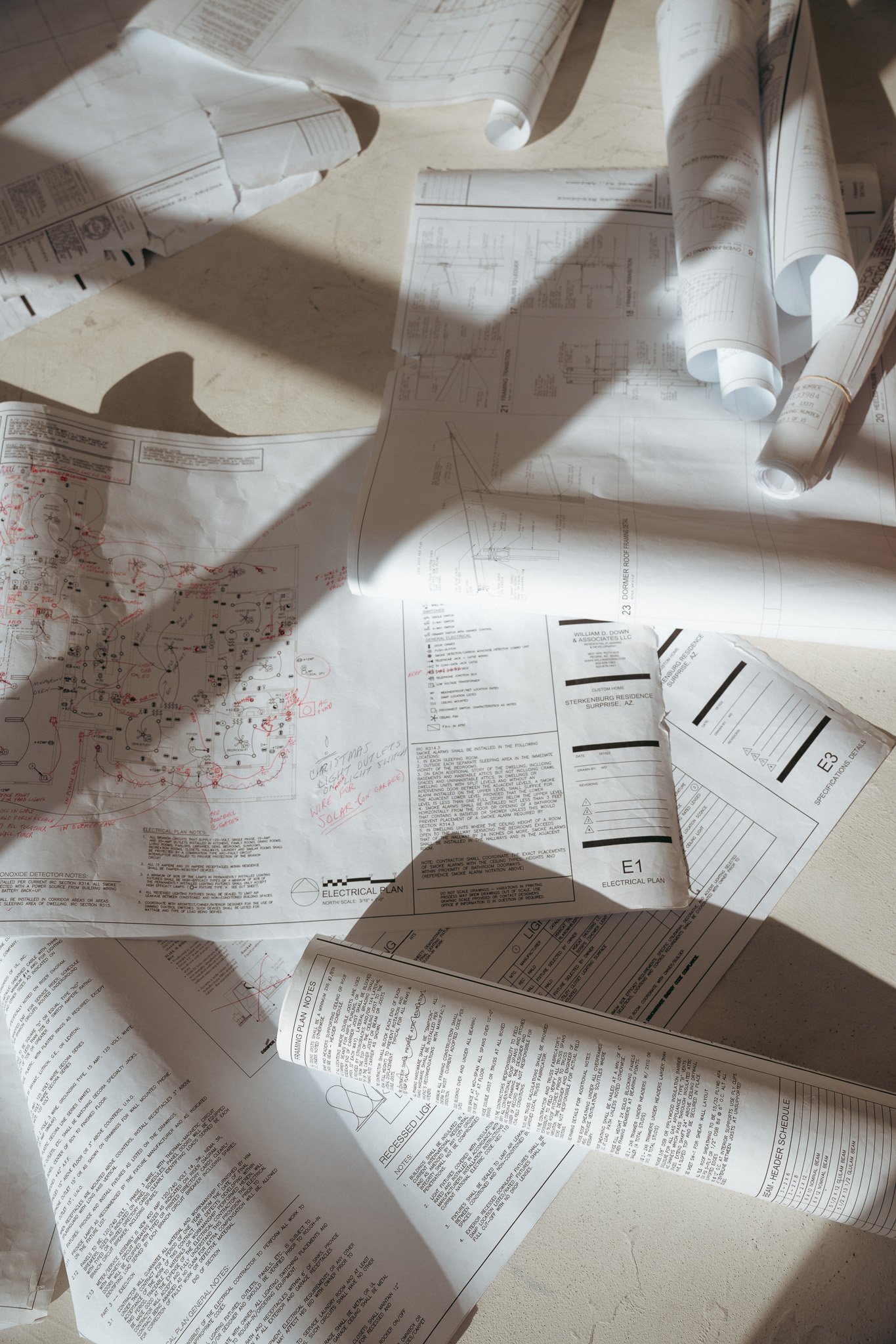 A scattered collection of architectural and electrical blueprints and plans, some sheet torn or rolled up, on a light-colored surface. Shadows of nearby objects cast across the papers.