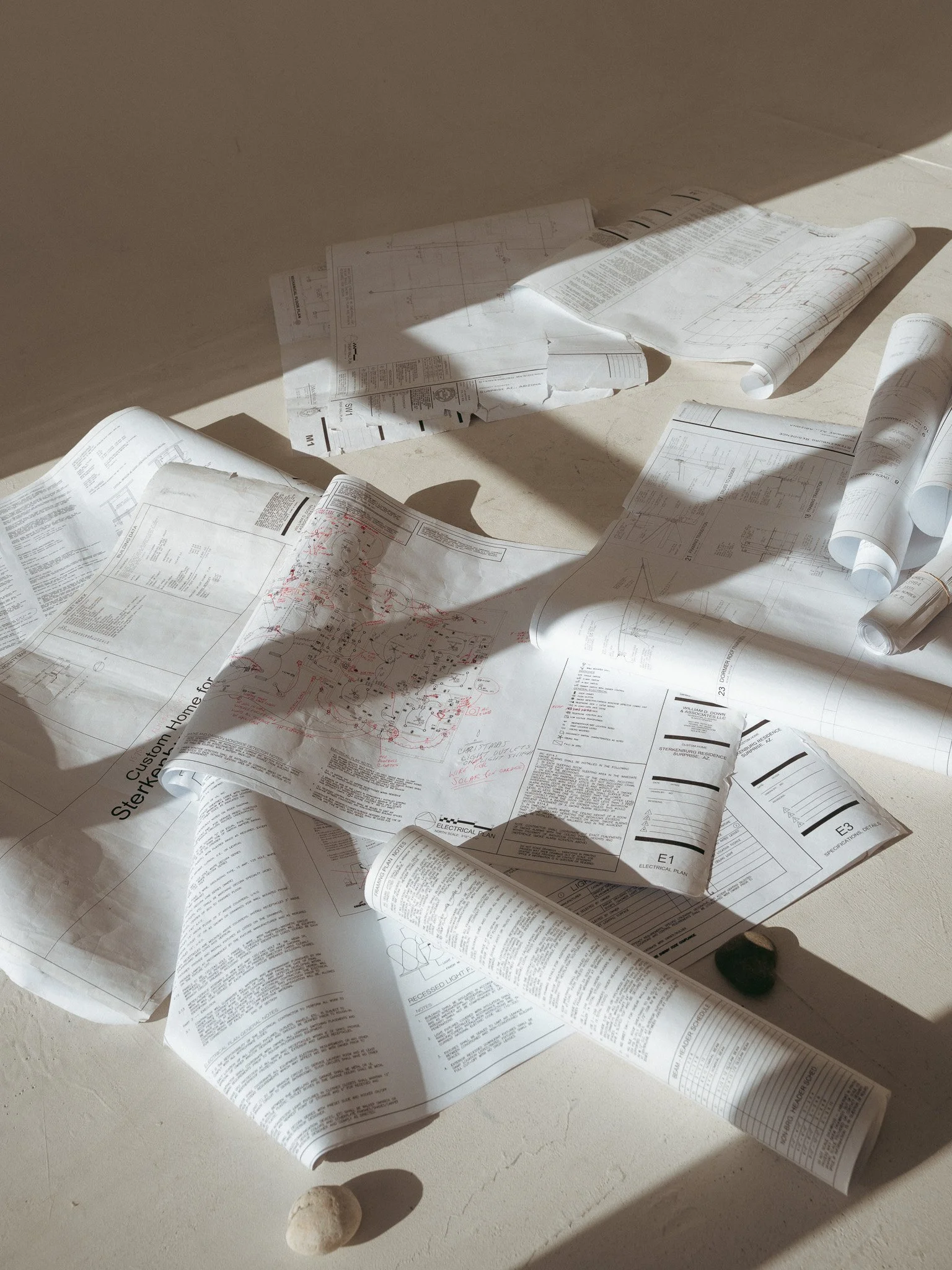 Scattered architectural blueprints and technical drawings on the floor with shadows cast across the papers.