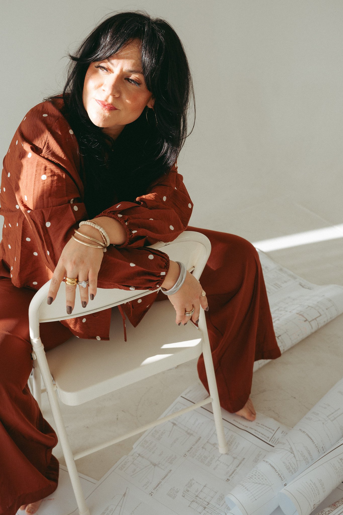 Woman with black hair wearing a red polka dot outfit sitting on a chair, pointing at architectural blueprints and plans on the floor, with more blueprints on the wall.