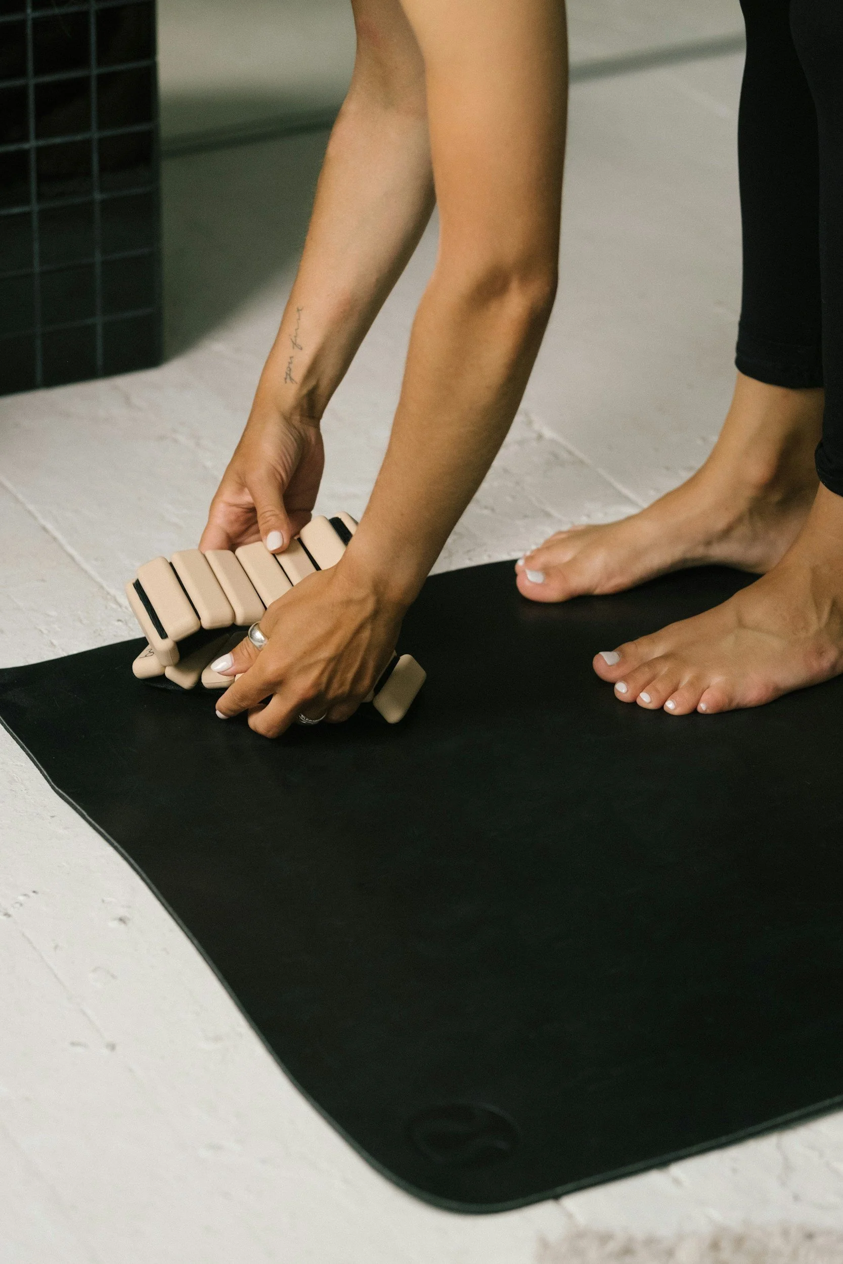 Close-up of a person's hands adjusting a beige yoga block on a black yoga mat, with a person barefooted next to them.