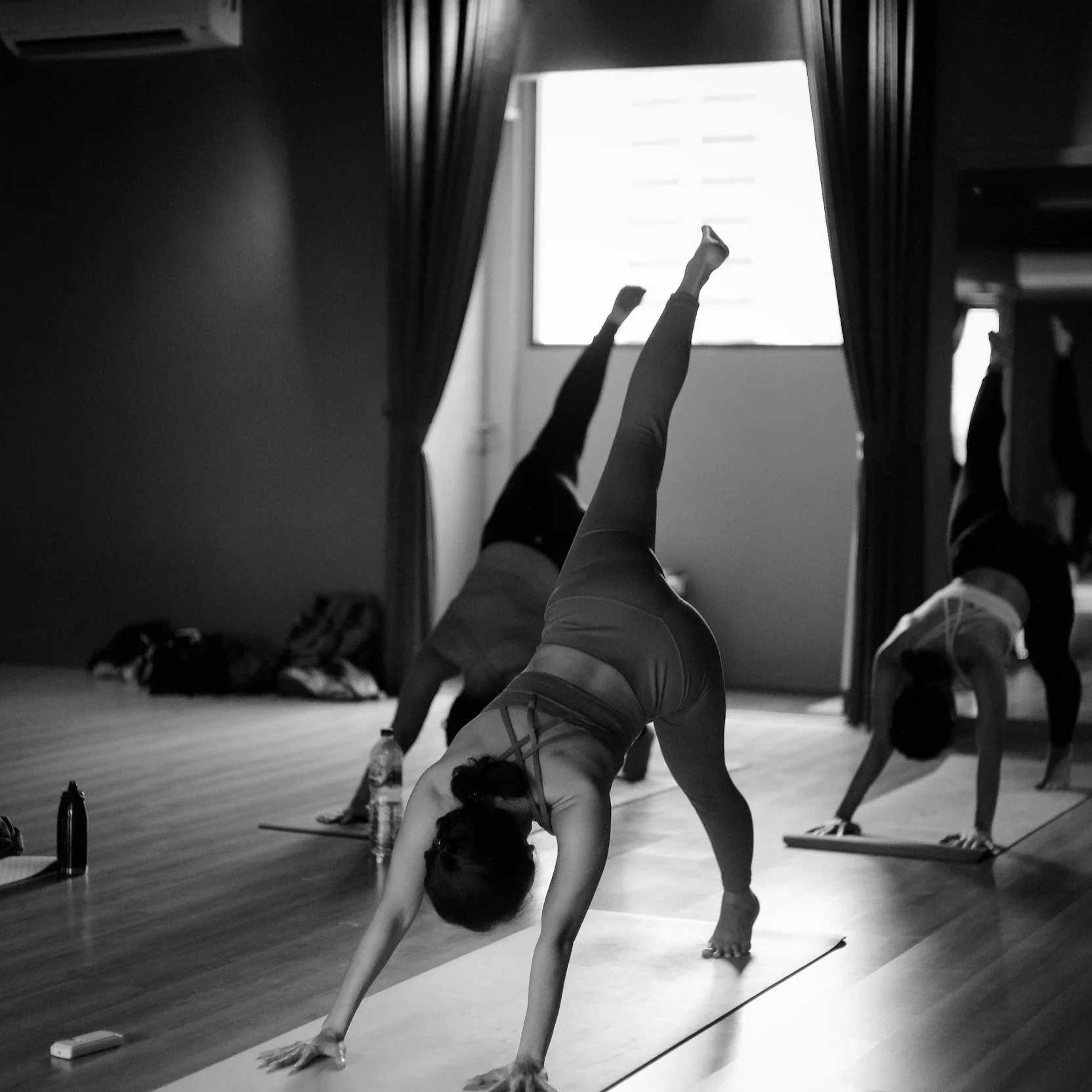 A woman in athletic wear practicing yoga, doing a handstand in front of a mirror in a studio with wooden flooring, with other people and items like water bottles and bottles in the background.
