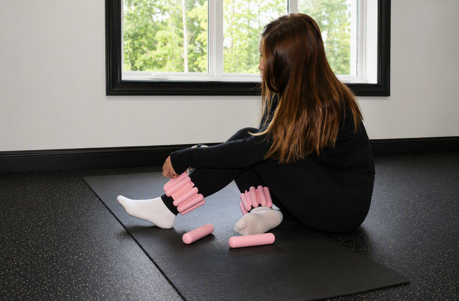 A woman seated on a black exercise mat in a room with large windows, stretching her legs with pink foam rollers around her calves and ankles.