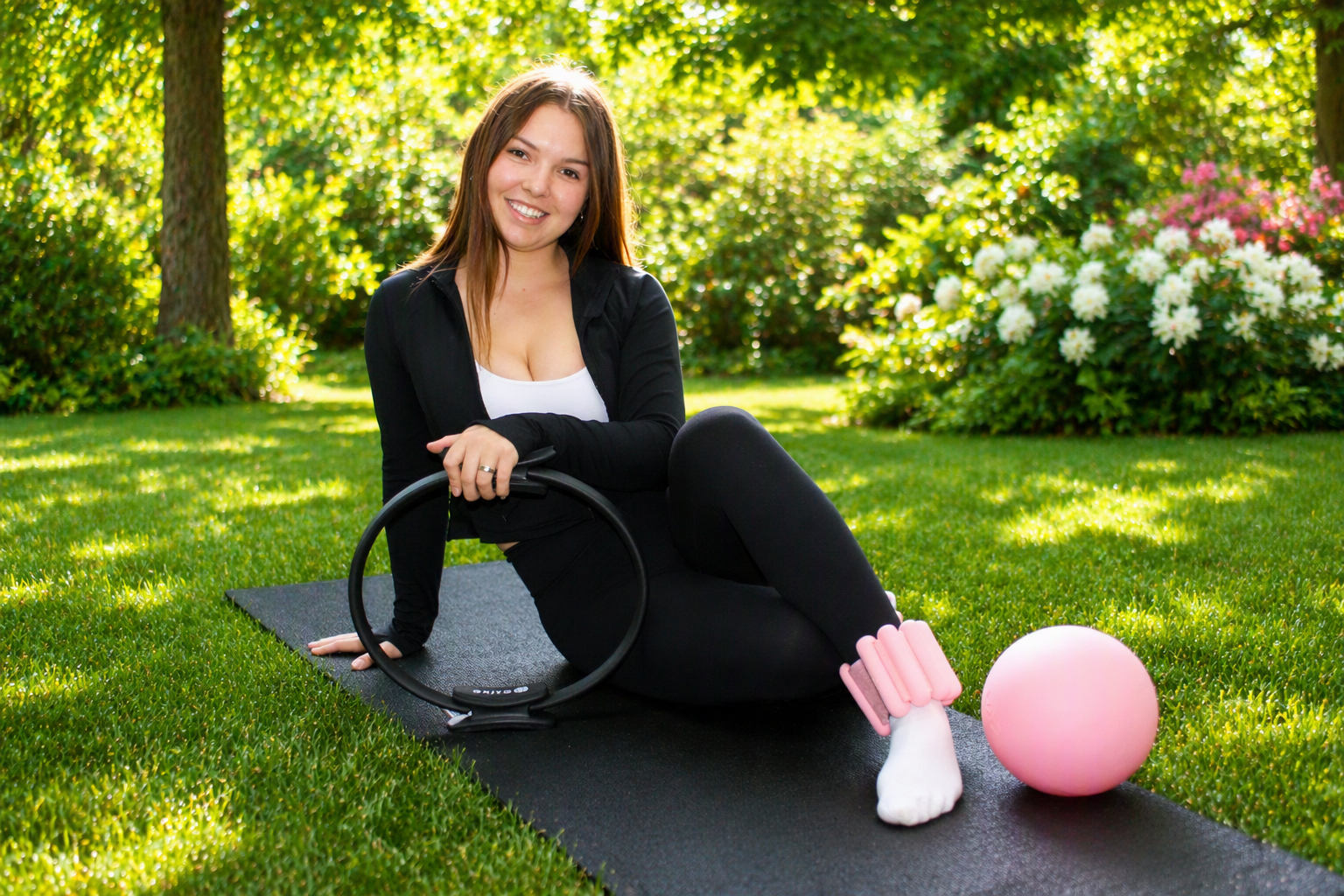 A woman sitting on a yoga mat outdoors in a park, holding a Pilates ring, surrounded by trees and flowering bushes on sunny day.