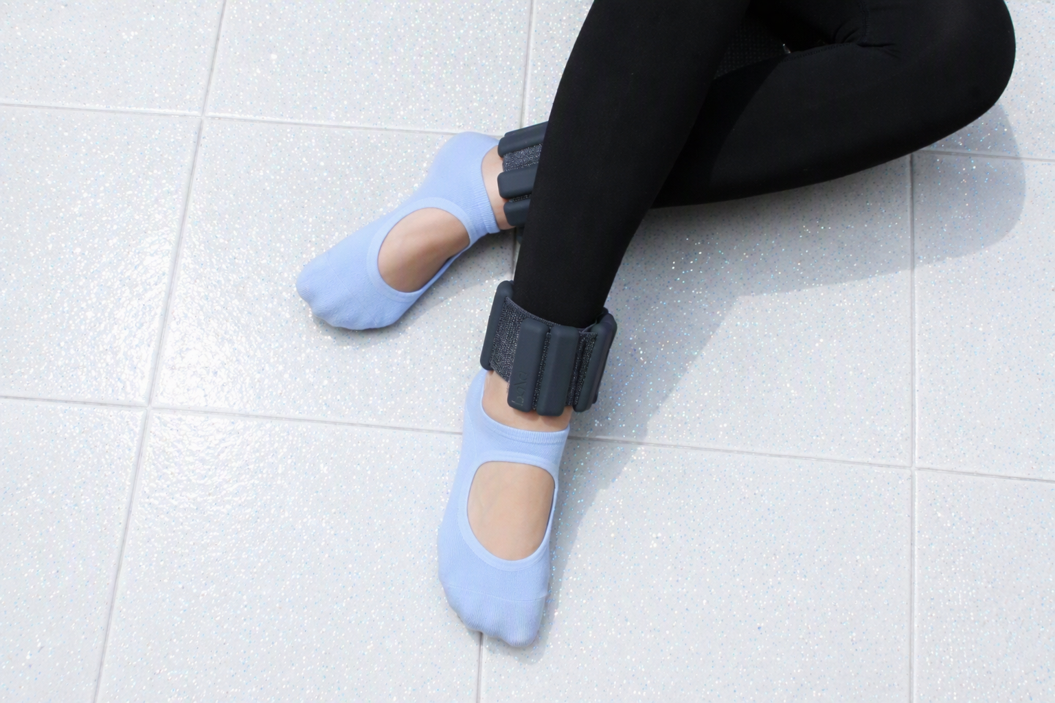 Person sitting on the floor wearing black pants and light blue socks with a gray ankle brace on each foot.