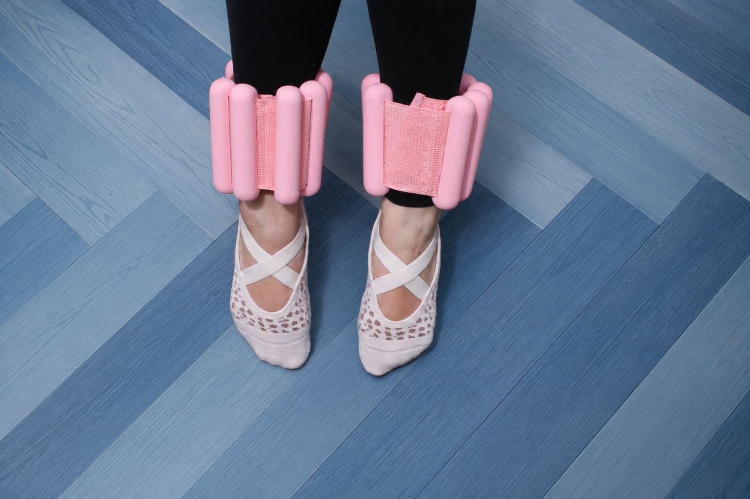 Person standing on blue wooden floor wearing ballet slippers and black leggings with pink foam ankle weights around both ankles.