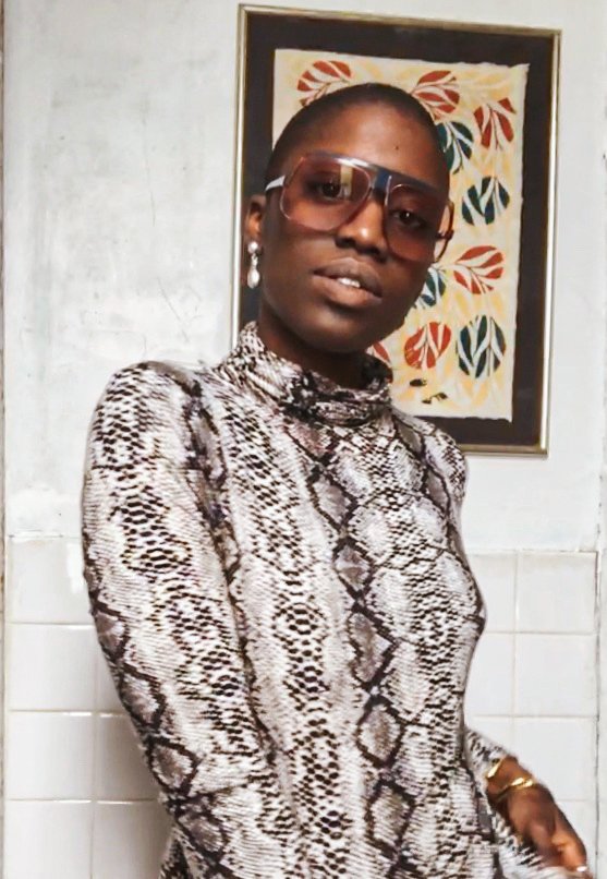 Woman wearing snake print top, large sunglasses, pearl earrings, and a gold bracelet, standing in front of a framed abstract art piece.