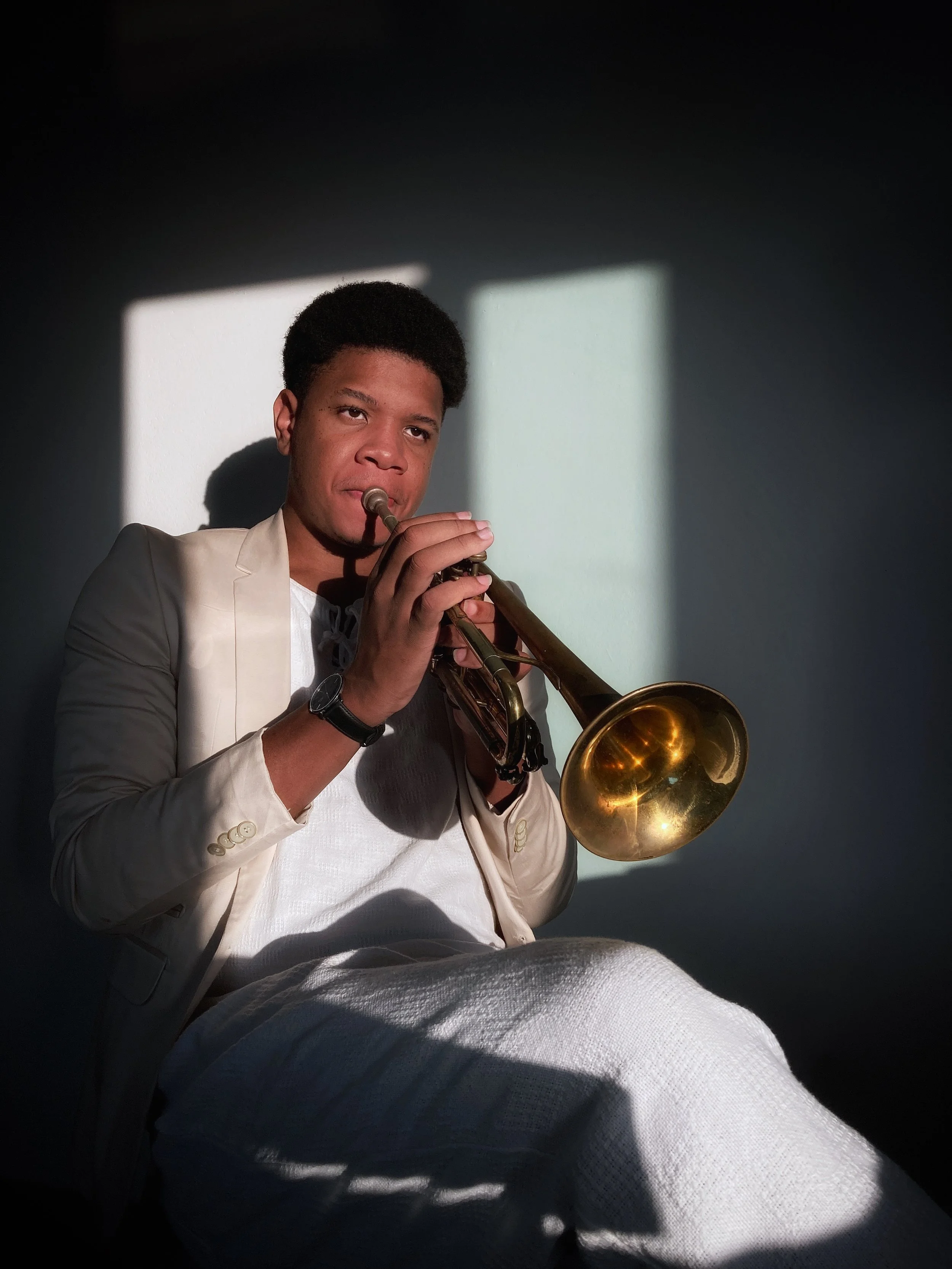 A young man playing a trumpet, wearing a white shirt and blazer, with sunlight casting shadows on a wall behind him.