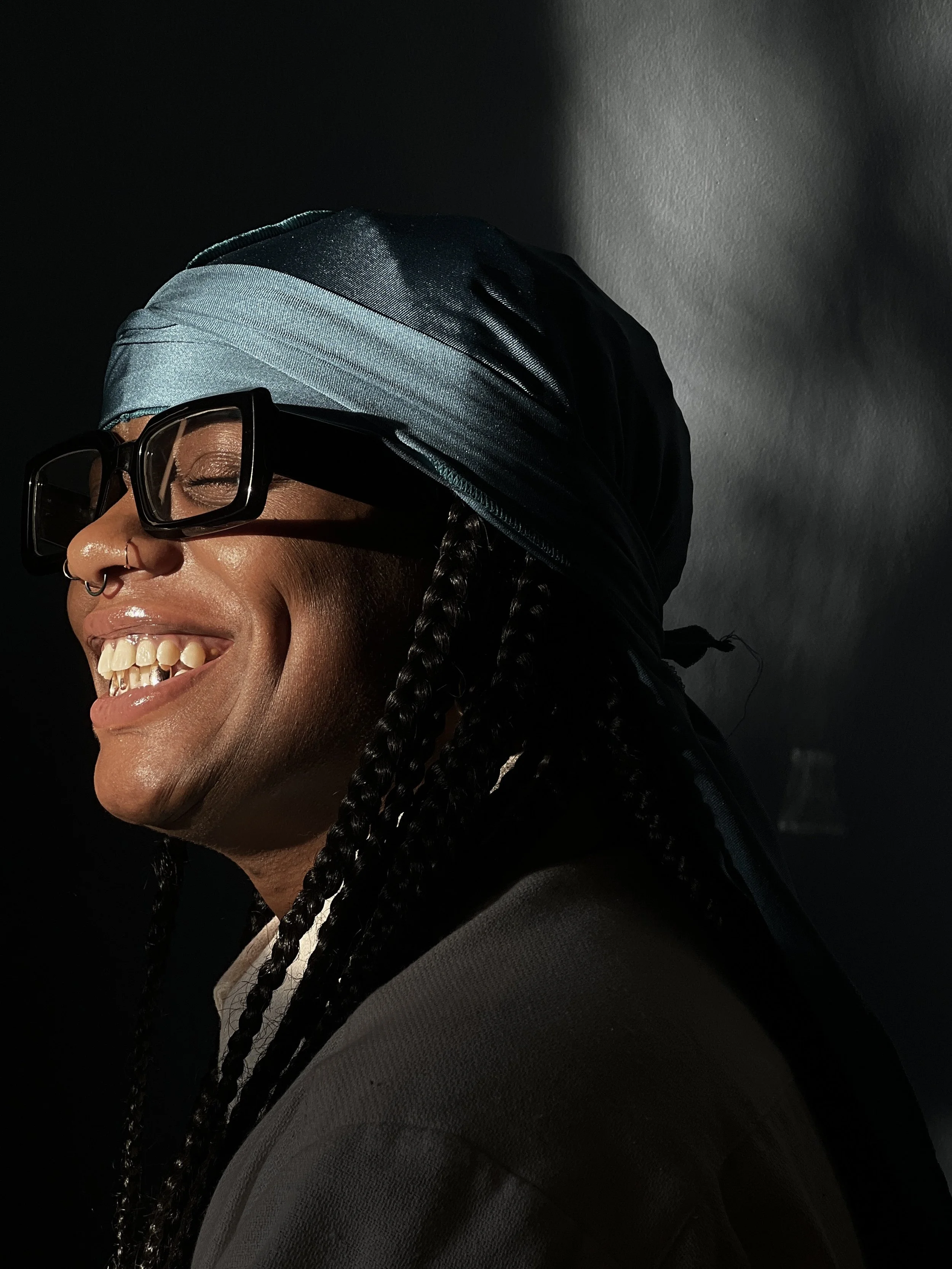 Close-up of a person with braided hair, wearing black sunglasses, a satin headscarf, earrings, and a nose ring, smiling against a dark background.