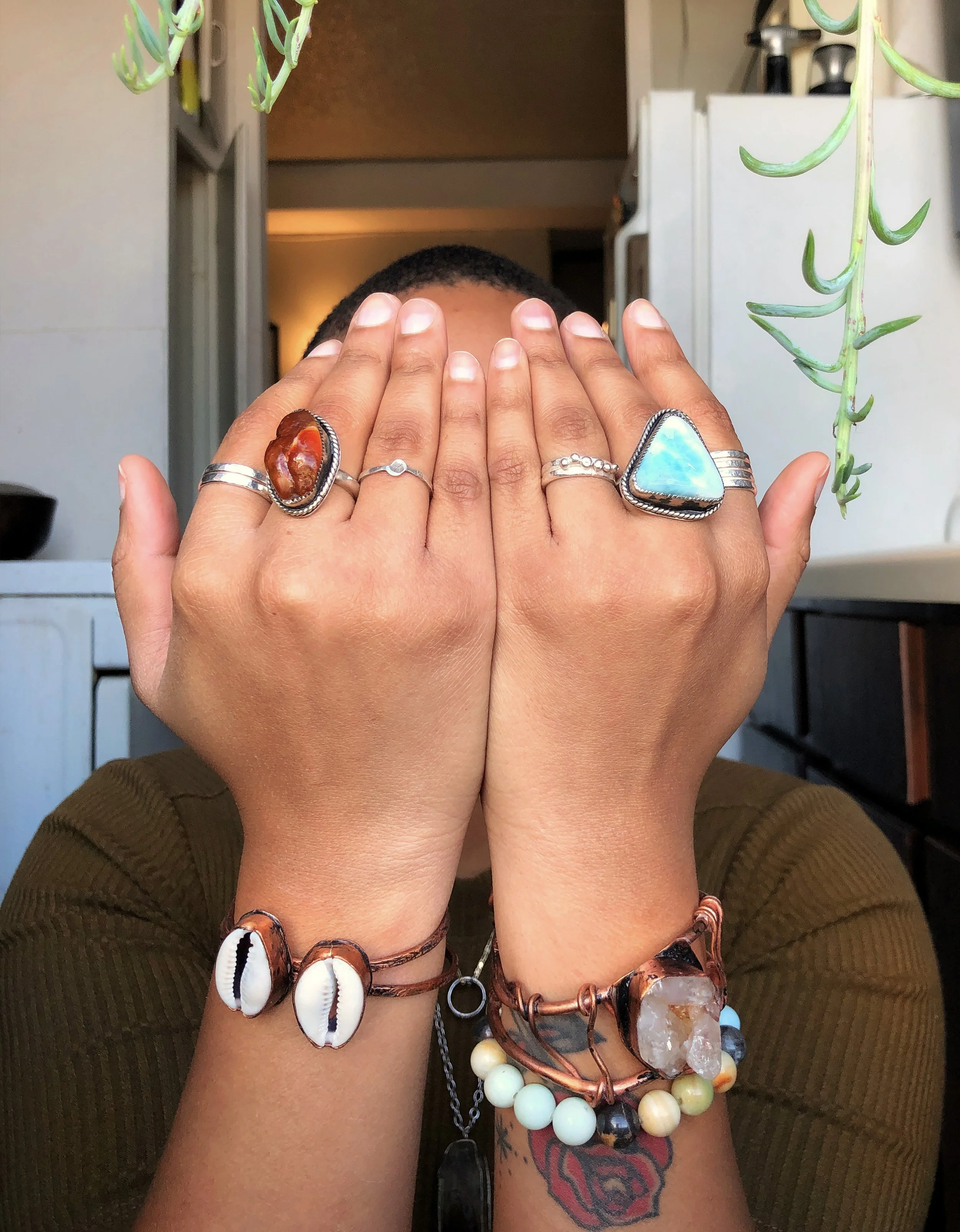 A person's hands covering their face, showing various jewelry including rings and bracelets with shells, stones, and beads, and a tattoo on the forearm, with a blurred background of indoor kitchen appliances and a hanging plant.