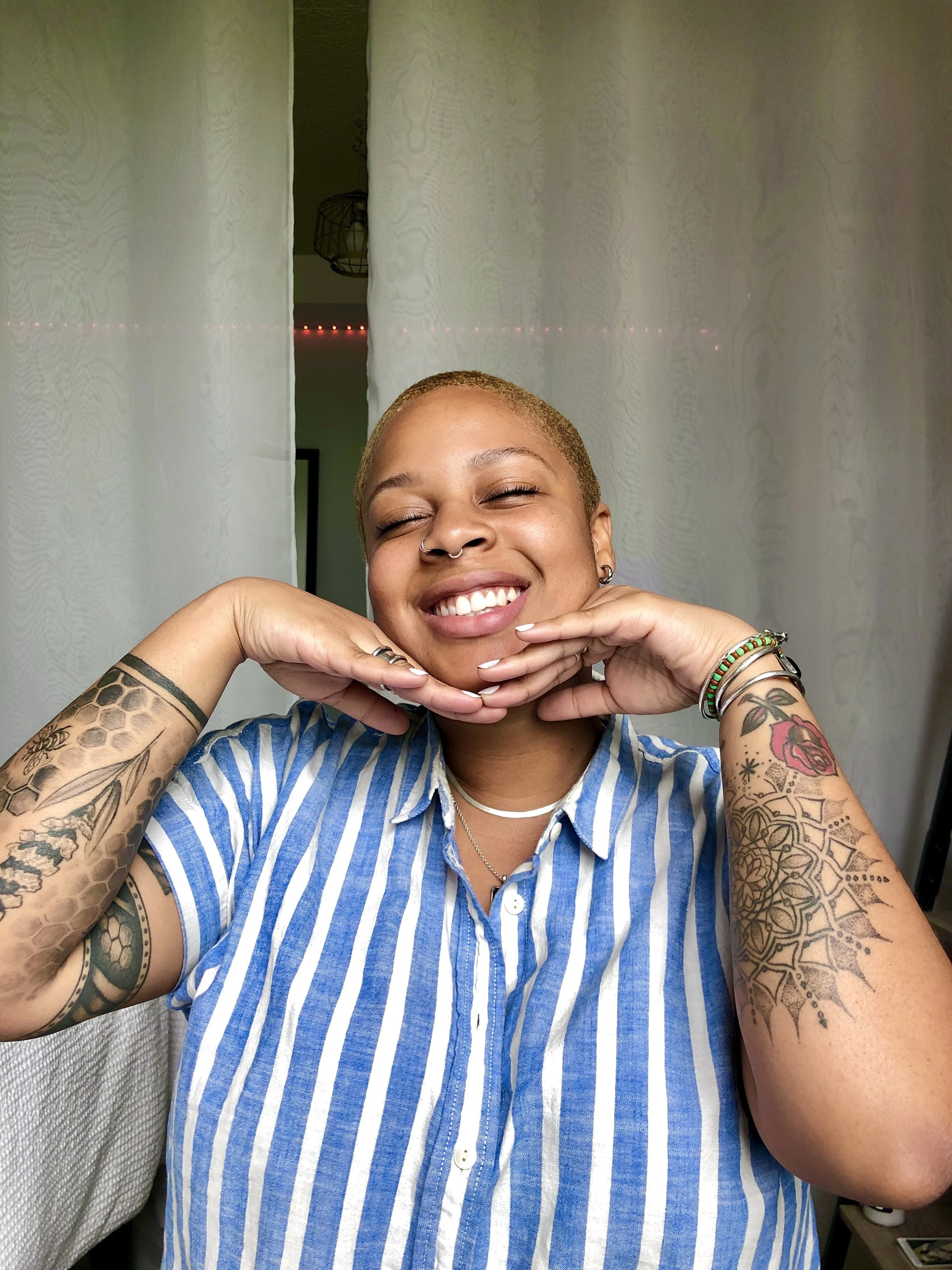 A person with short blonde hair and tattoos on both arms, smiling with eyes closed, holding their chin with both hands, standing in front of white curtains, wearing a blue and white striped shirt.