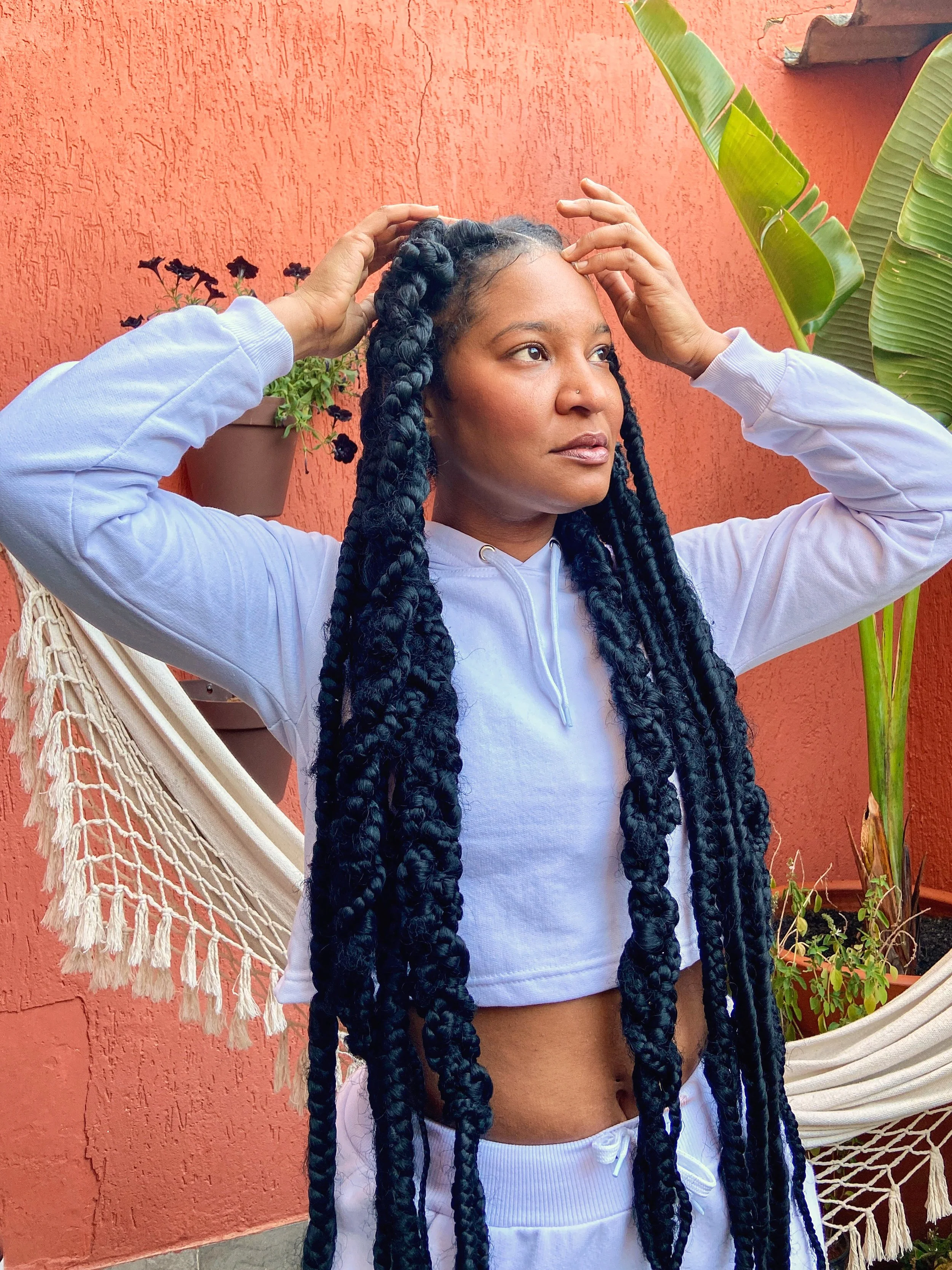 A woman with long black rope twists in her hair, wearing a white hoodie, standing in front of a red textured wall with plants, adjusting her hair with both hands.