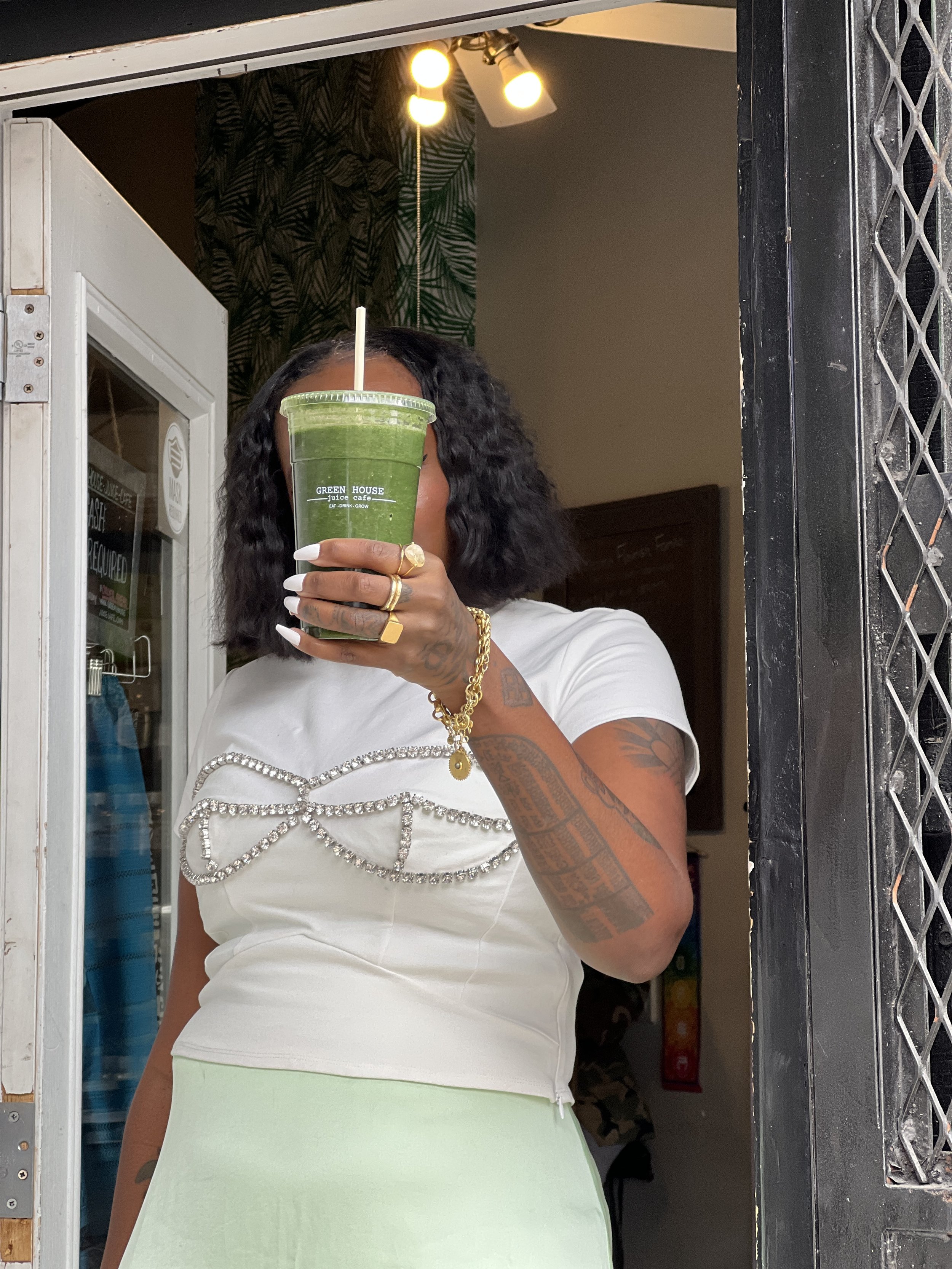A woman with dark, curly hair and tattoos on her arm is holding a green smoothie from Green House Juice Cafe in front of her face, obscuring her features. She is wearing a white top with rhinestone embellishments and a light green skirt, and is stand