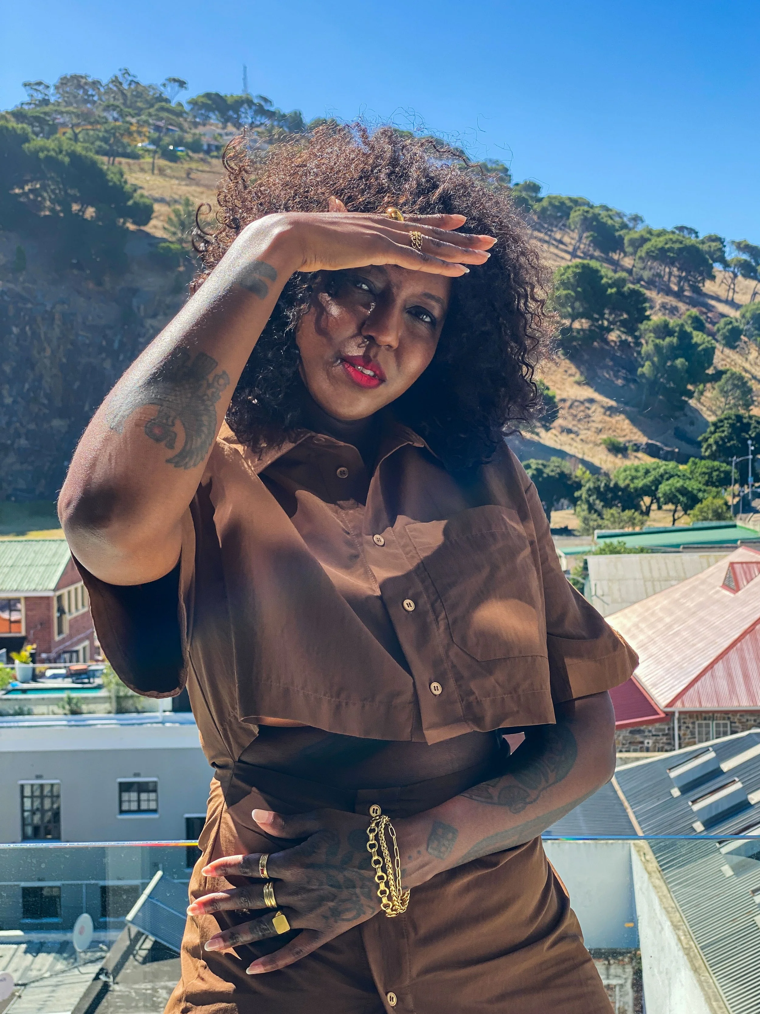 A woman with curly hair and tattoos on her arm, wearing rings, a gold chain, and a brown shirt, saluting or shielding her eyes from the sun, standing outdoors with a mountainous landscape and buildings in the background under a clear blue sky.