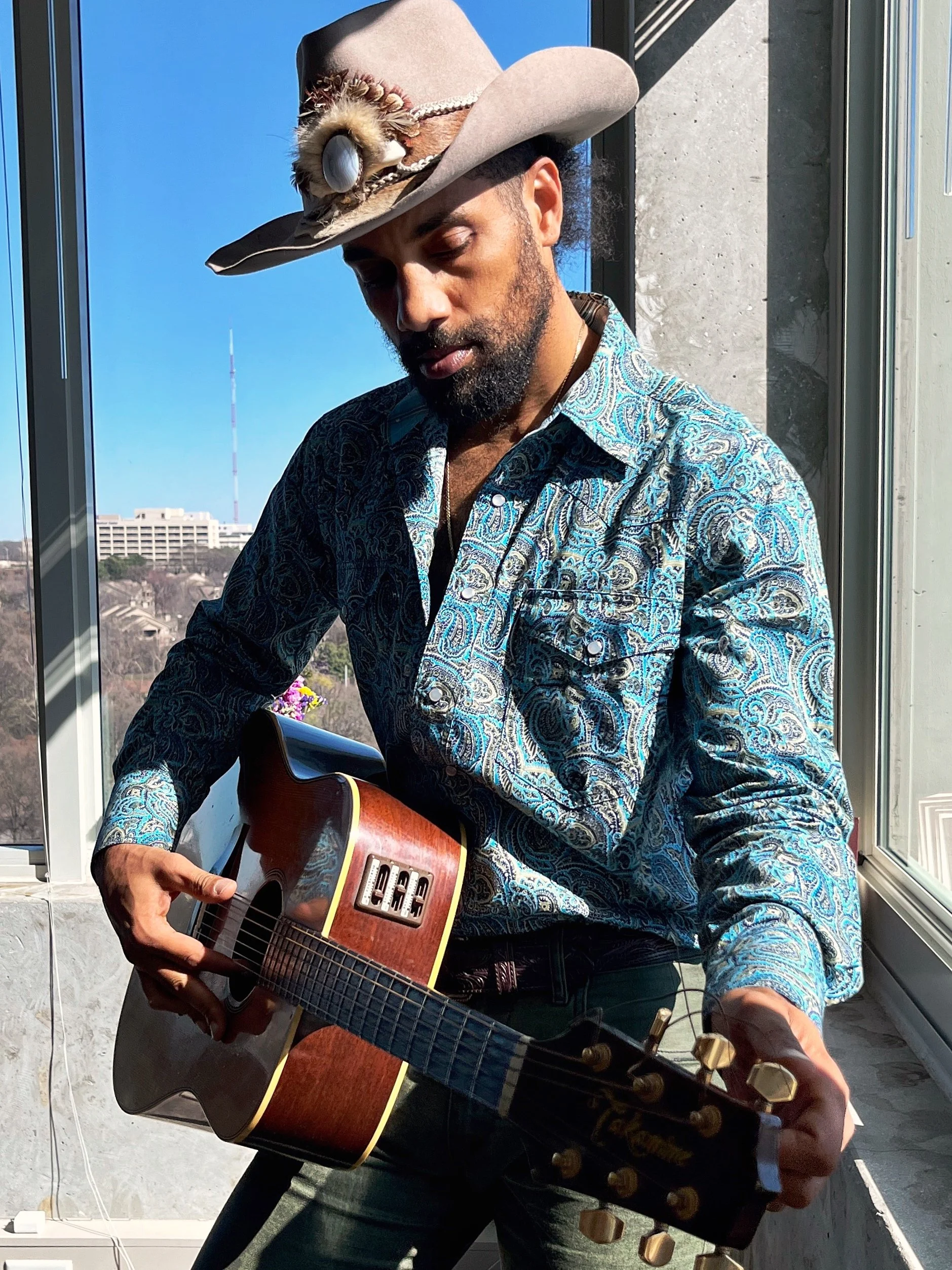 A man wearing a wide-brimmed beige hat adorned with feathers and a pearl necklace, playing an acoustic guitar with a scenic city view in the background.