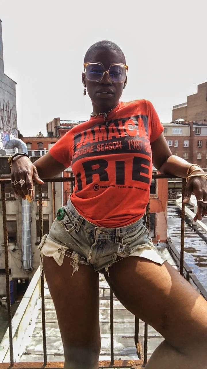 A confident woman with short hair and sunglasses standing on a rooftop with urban buildings in the background, wearing a red graphic t-shirt and torn denim shorts.