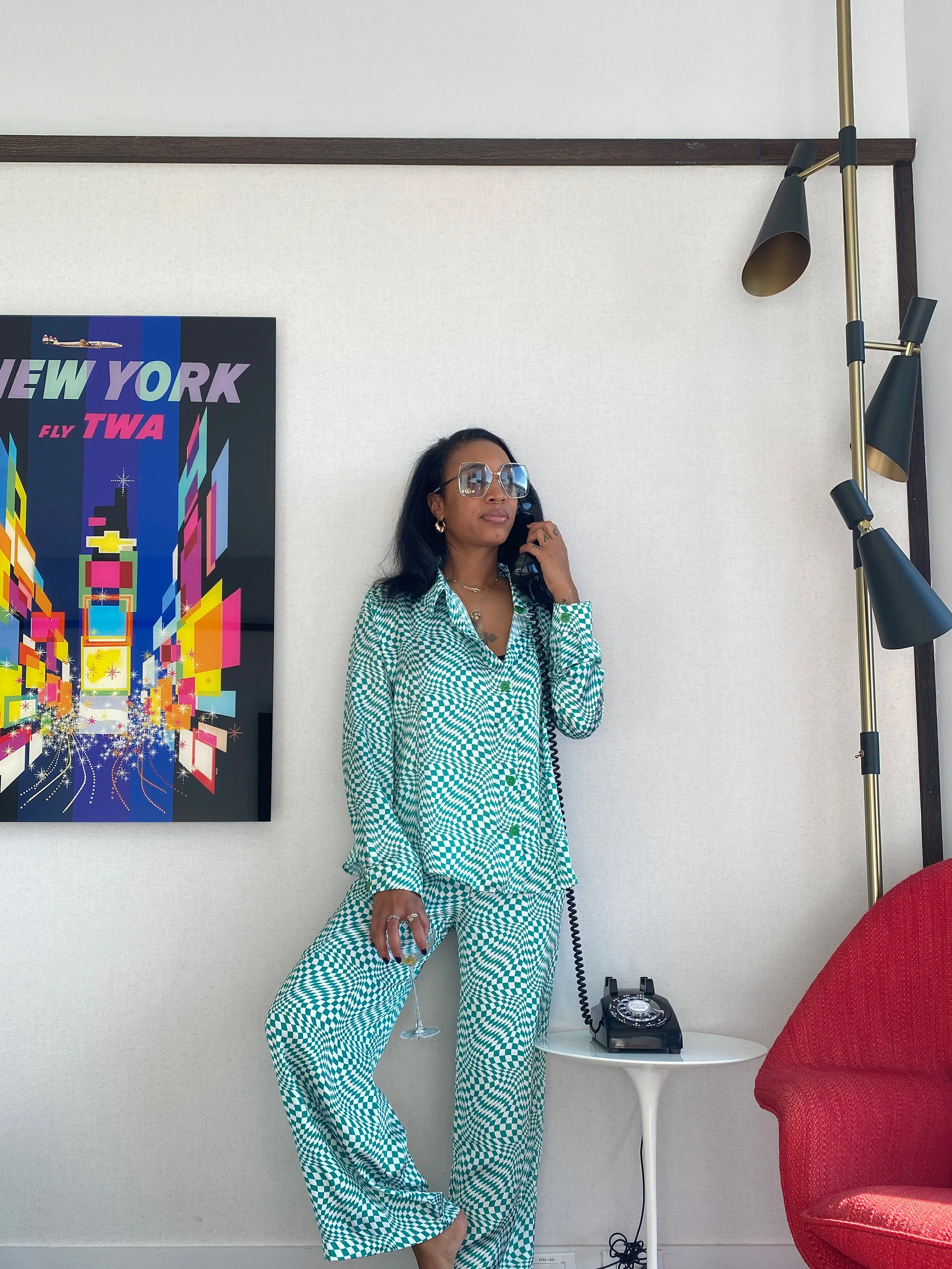 A woman with long dark hair, wearing oversized sunglasses and a green and white checkered pajama set, is standing in a furnished room, holding a vintage rotary phone and a wine glass with a clear beverage, appearing relaxed.