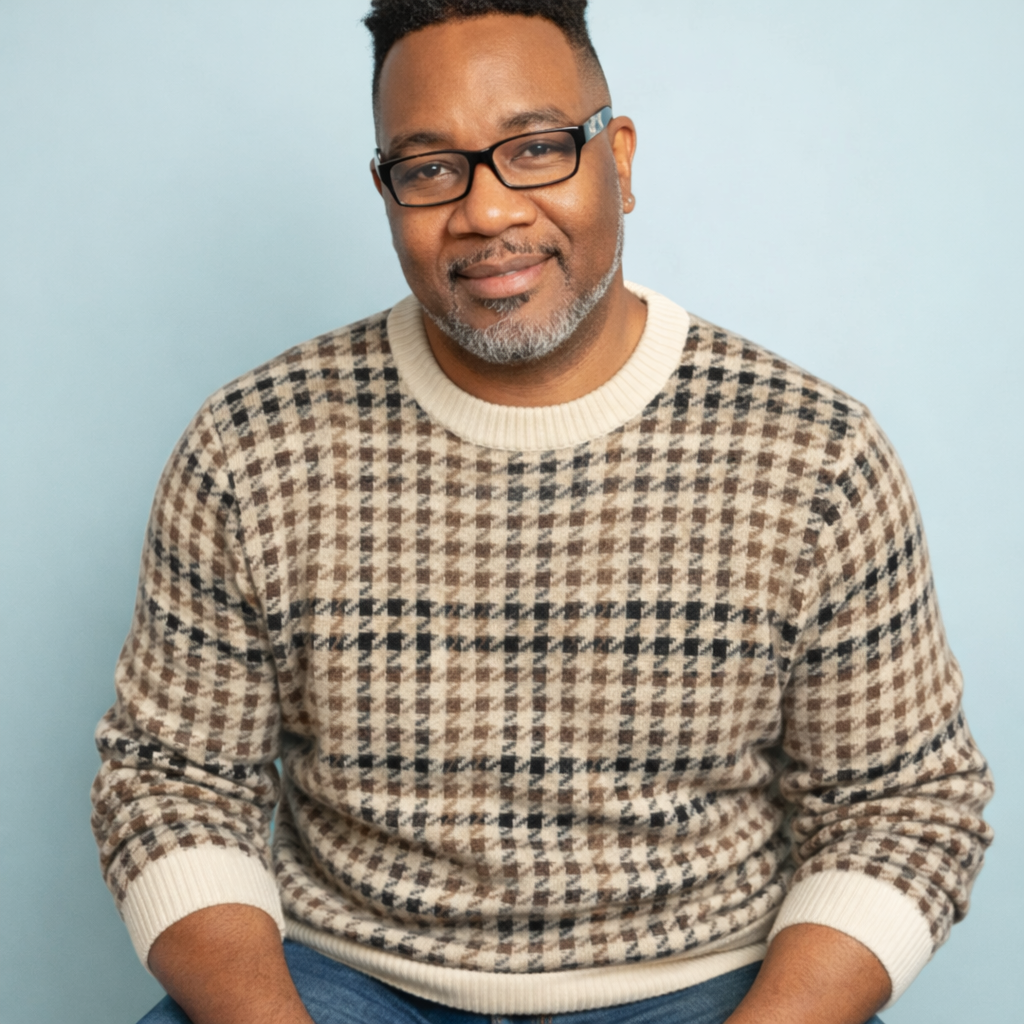 A middle-aged man with glasses, gray beard, wearing a beige and brown houndstooth sweater and blue jeans, smiling against a light blue background.