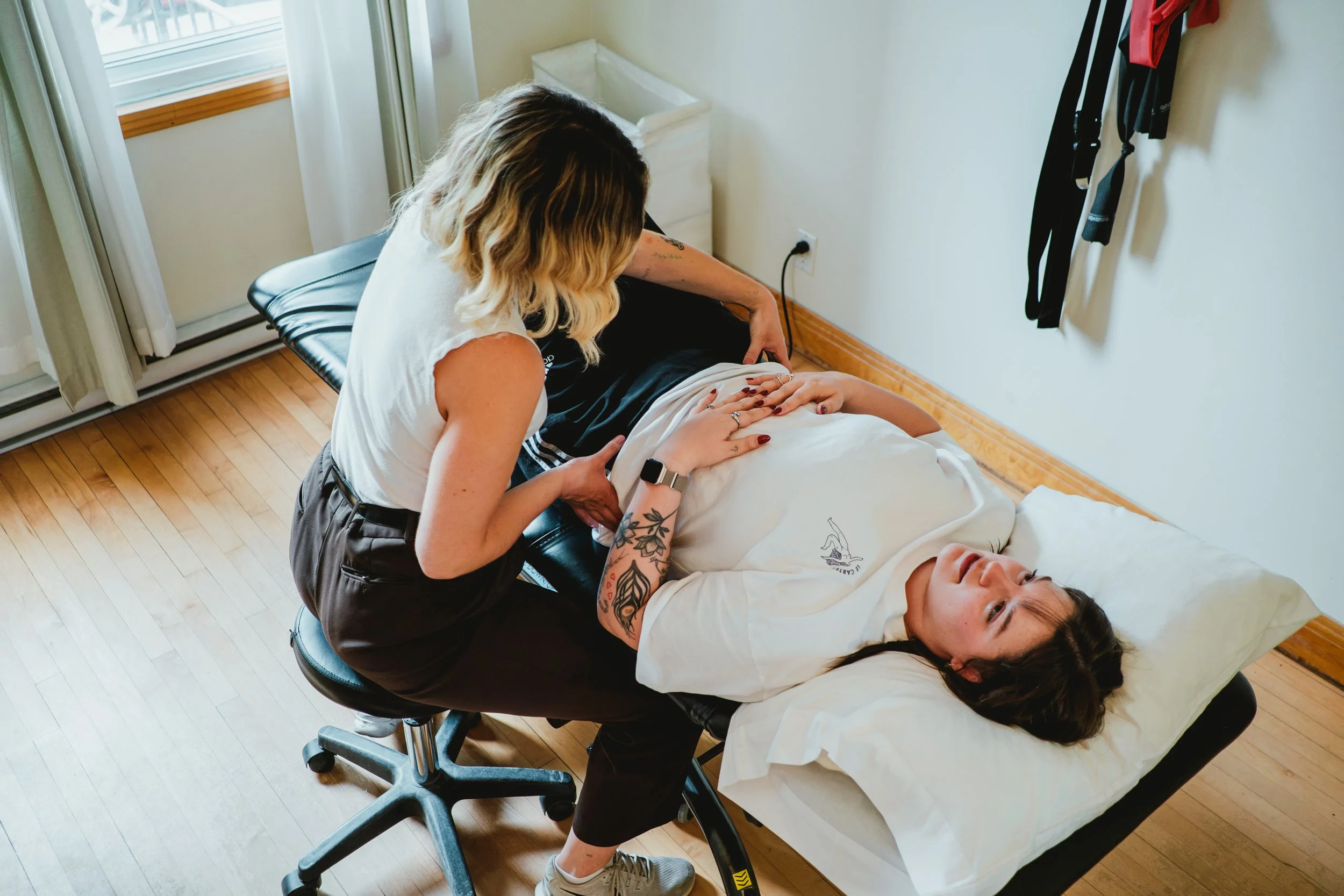 Une femme en cours de massage d'une autre personne allongée sur une table, dans une pièce lumineuse avec un sol en bois, une fenêtre avec des rideaux et un panier à linge.