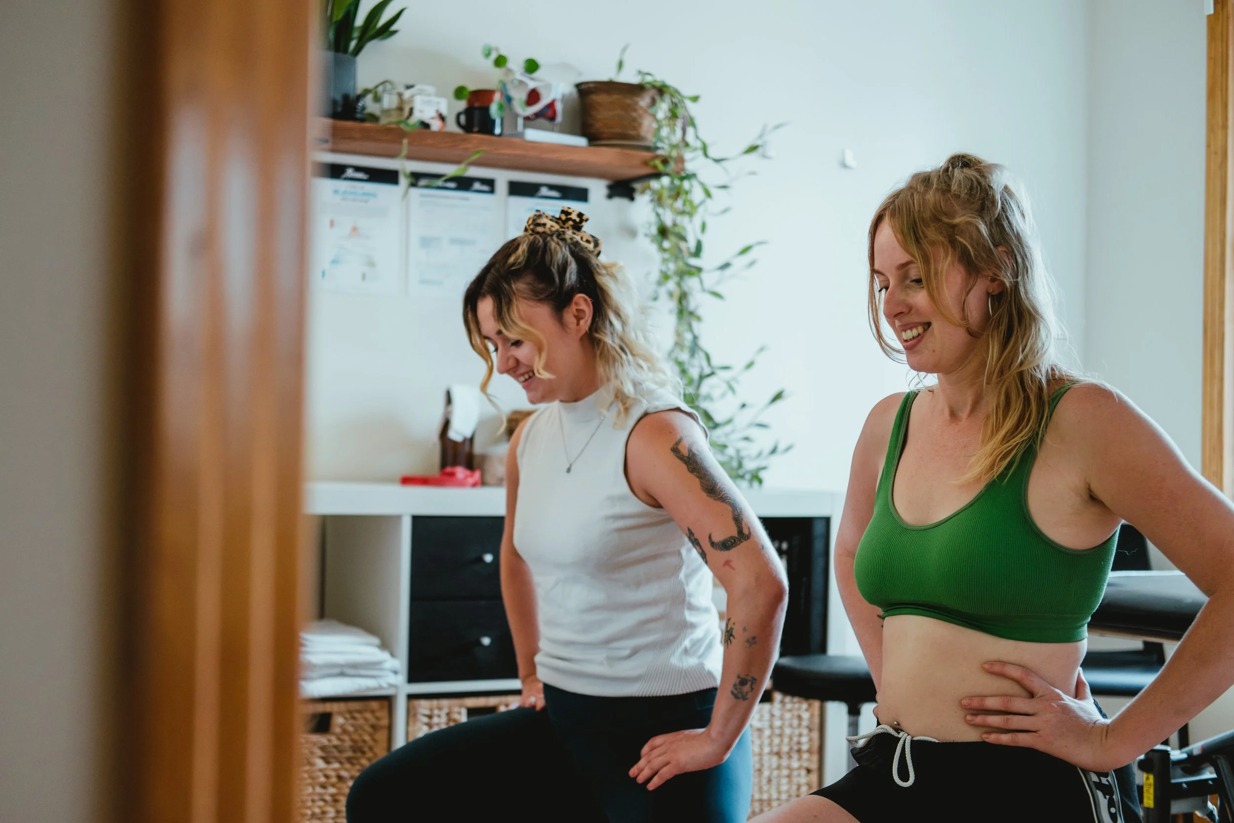 Deux jeunes femmes souriantes participent à une séance de yoga à l'intérieur, l'une porte un t-shirt blanc et la autre une brassière verte, avec un décor intérieur comprenant des plantes et des dossiers médicaux accrocher au mur.