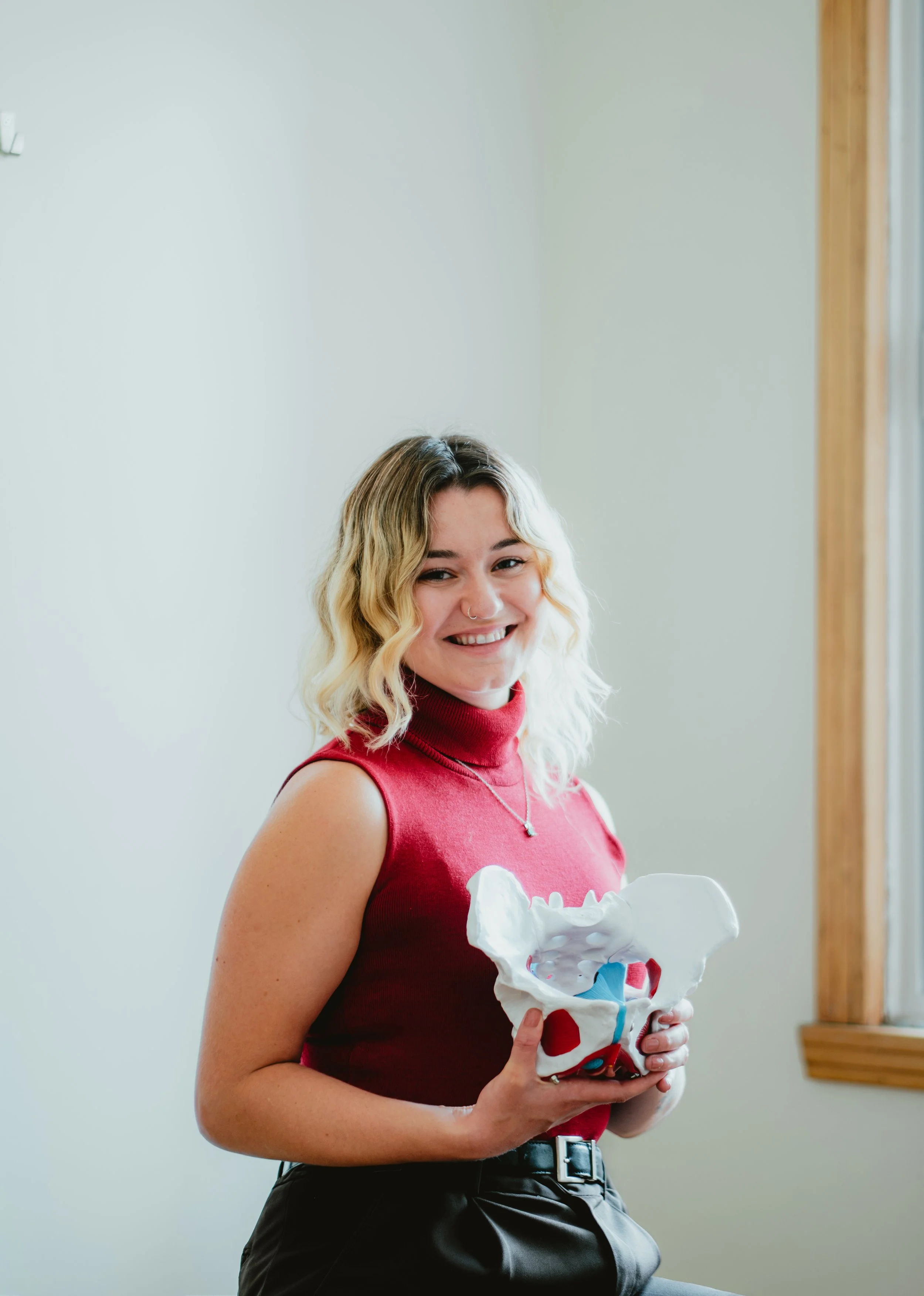 Une femme souriante tenant un modèle du squelette humain, portant un haut sans manches rouge, dans une pièce avec mur blanc et fenêtre avec cadre en bois.