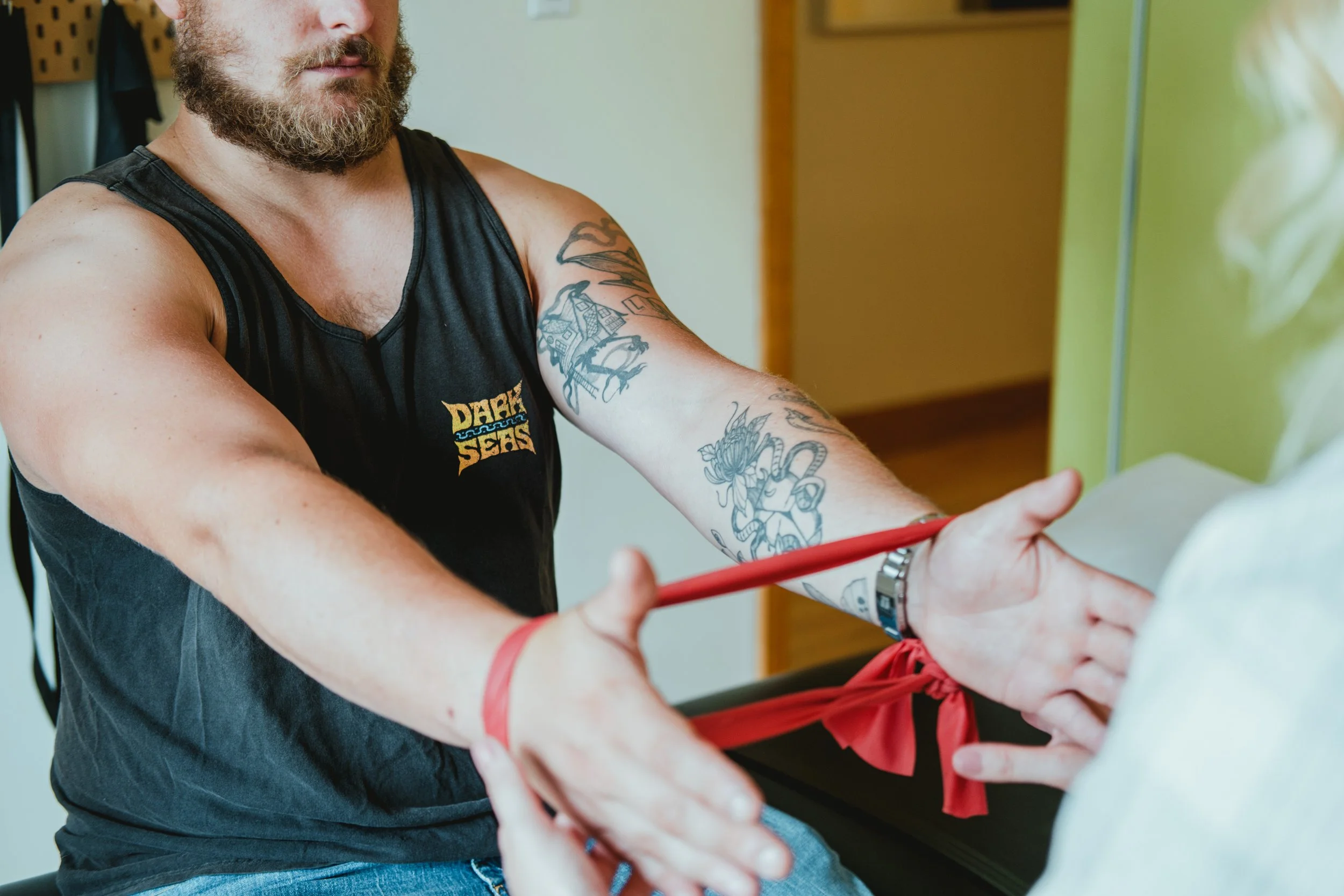 Un homme avec une barbe et des tatouages sur le bras, portant un débardeur noir, s'entraîne avec une bande de résistance rouge, en salle de sport.