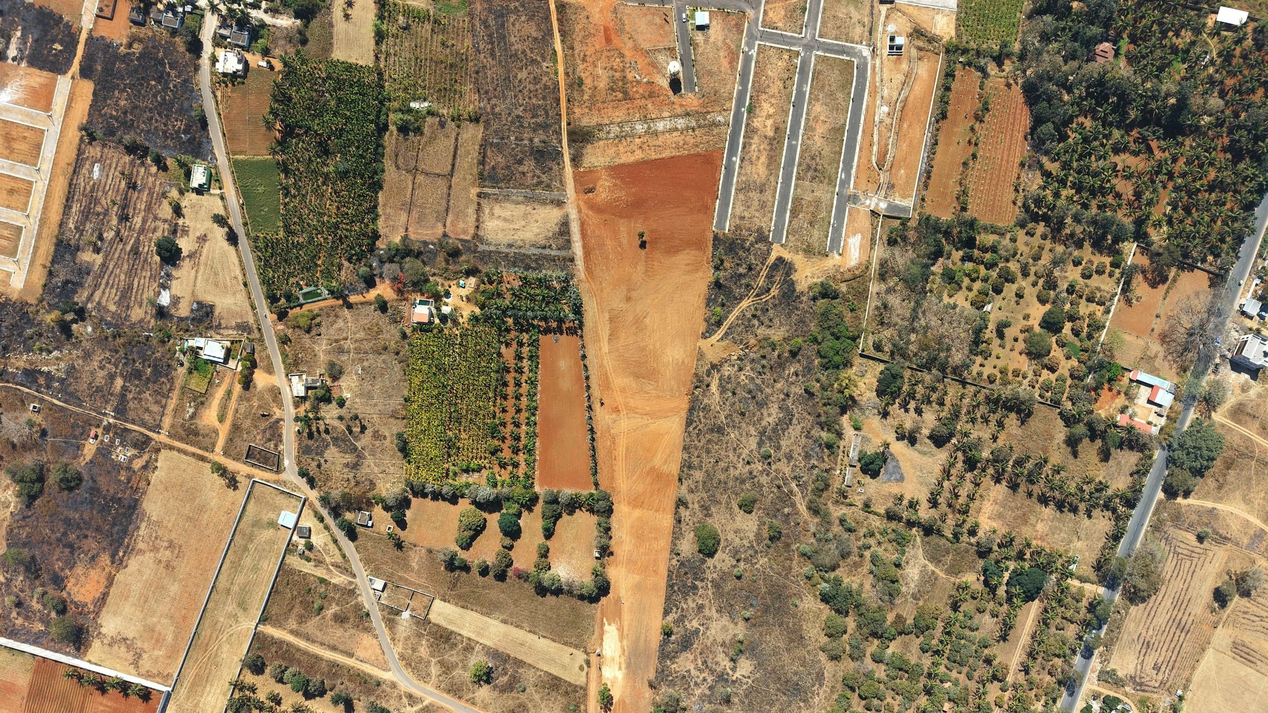 Aerial view of farmland with fields, trees, and roads. Some fields are brown, others are green or cultivated.