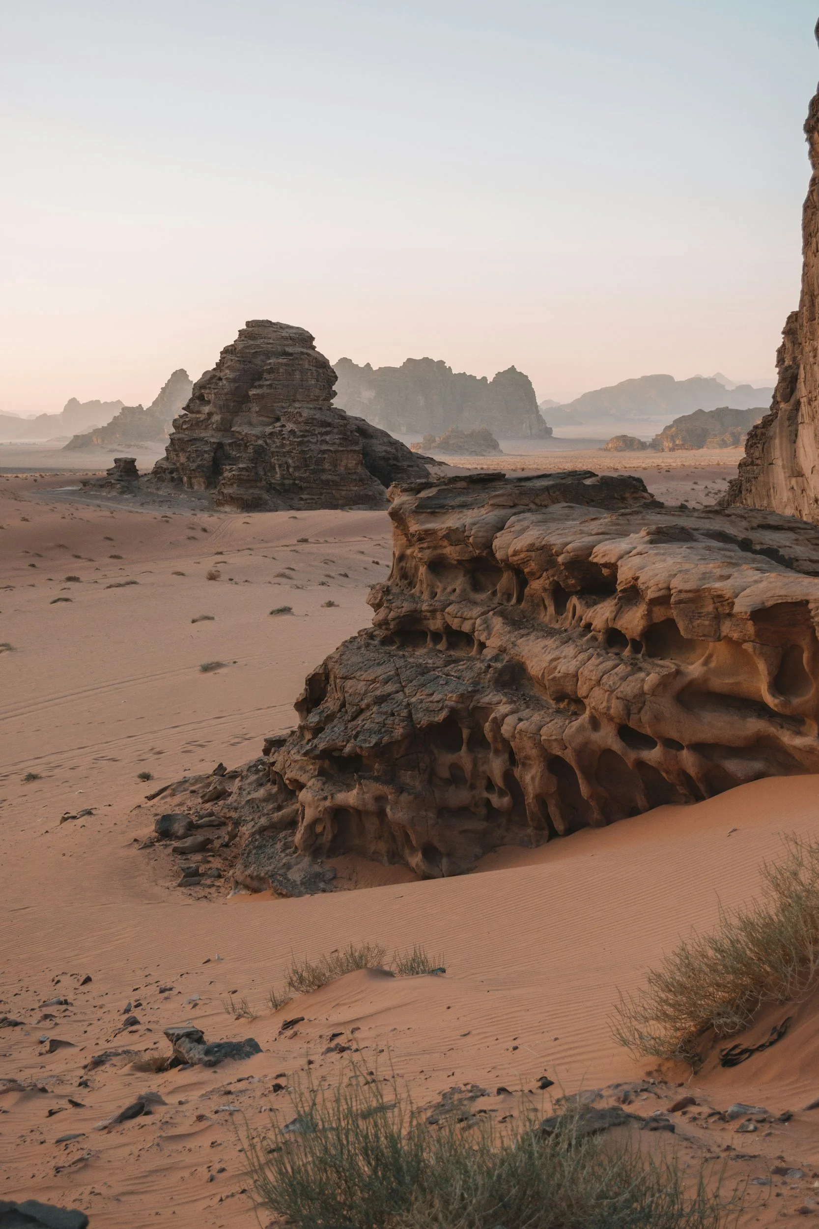 A desert with large rock formations and sand dunes, sparse vegetation, and a hazy sky.