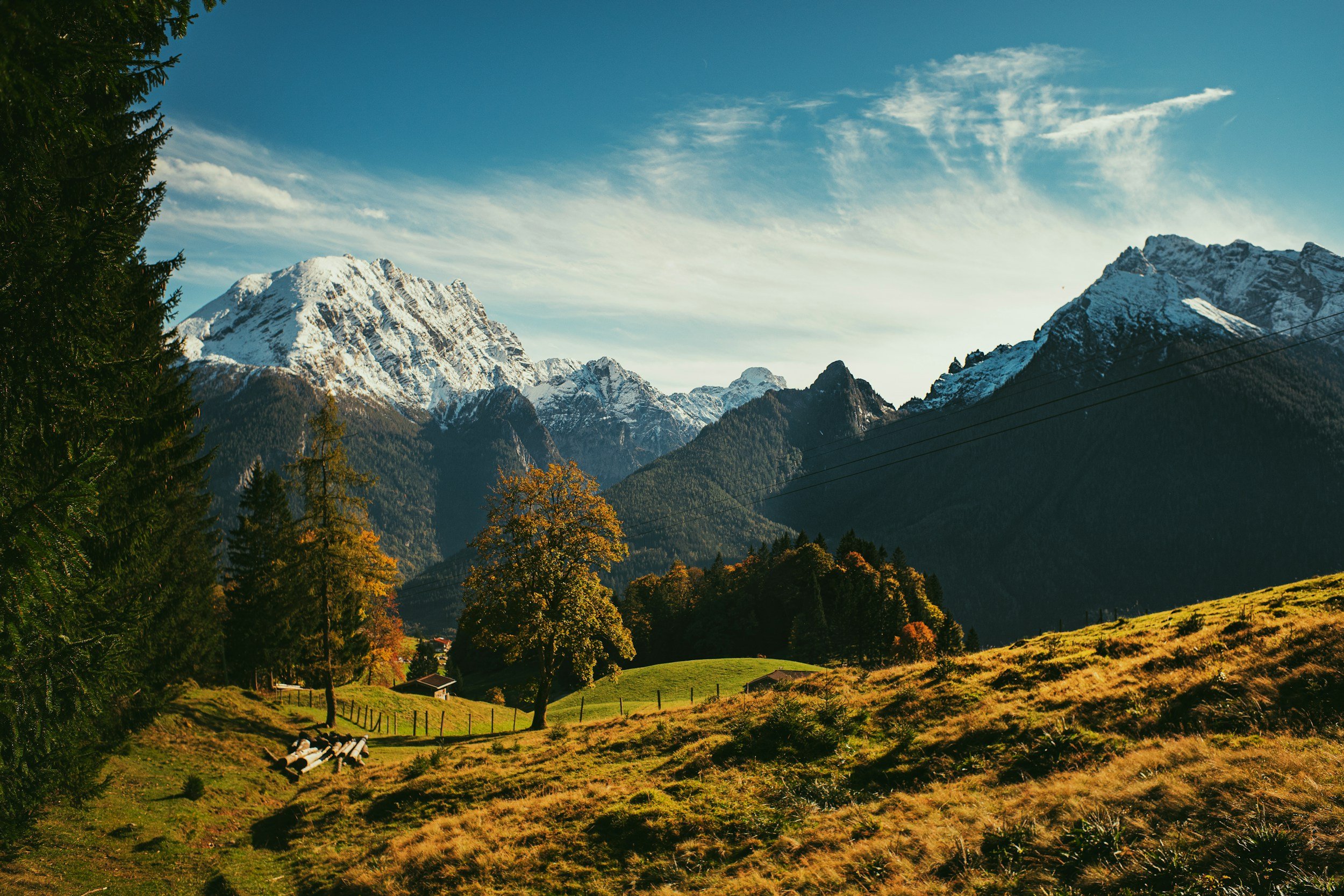 Snow-capped mountains in the background with a green valley and autumn trees in the foreground under a partly cloudy sky.