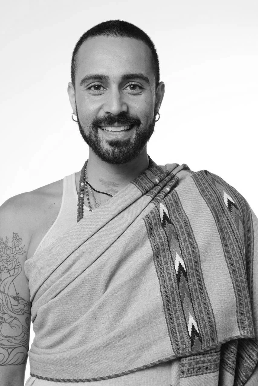 A smiling man with short hair and a beard, wearing earrings, necklaces, a sleeveless shirt, and a traditional patterned shawl draped over one shoulder.