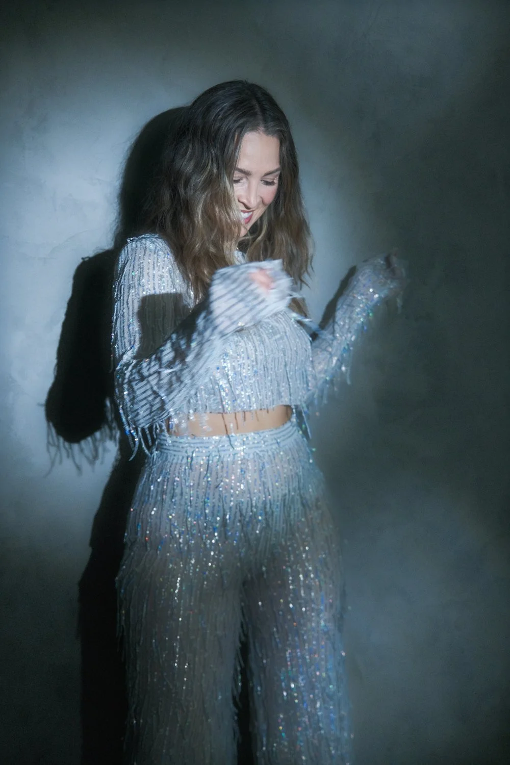 A woman with long, wavy hair smiling and looking down, wearing a sparkling silver two-piece outfit with sequins and fringe, standing against a plain gray wall.