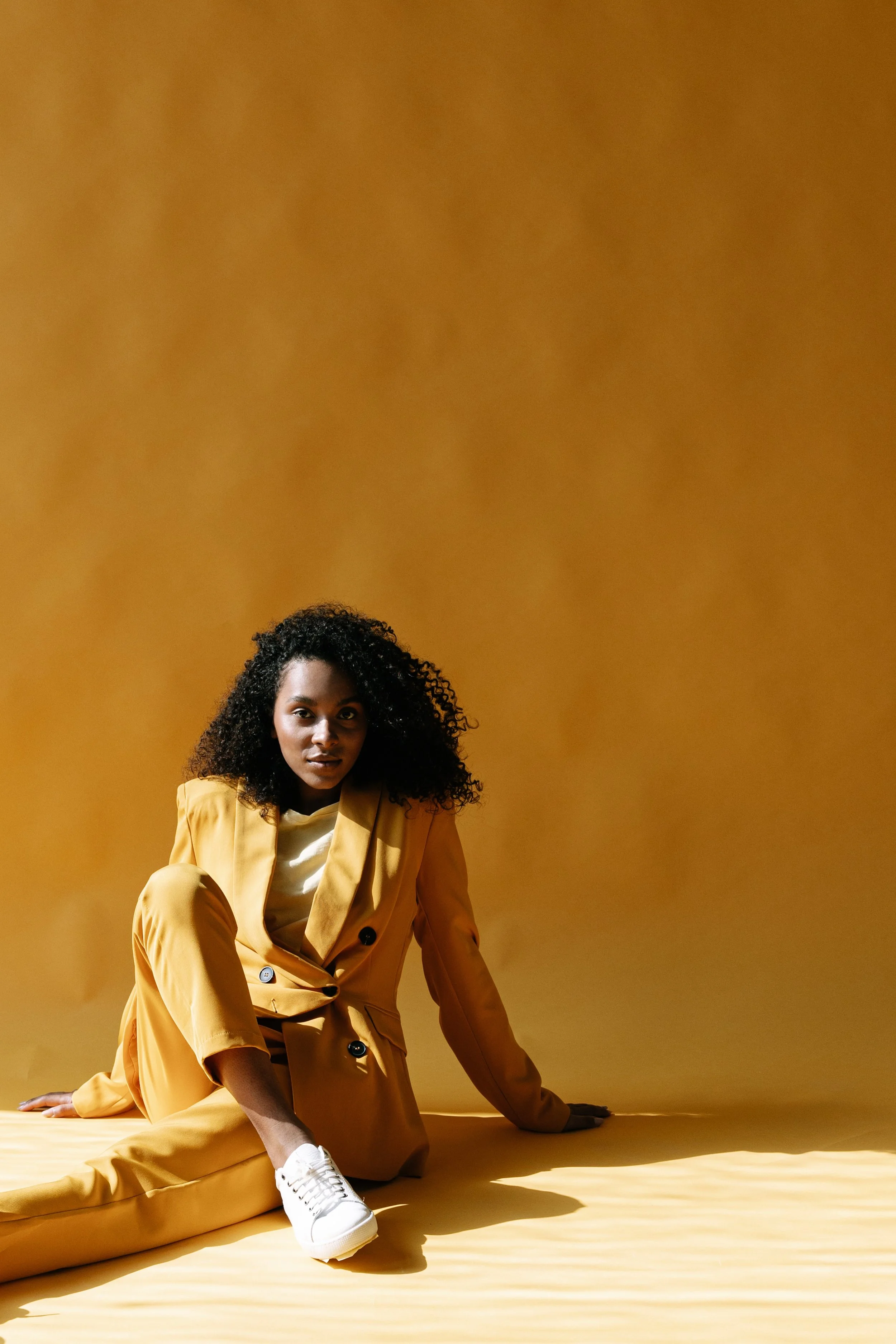 A woman with curly black hair wearing a yellow suit and white sneakers is sitting on a yellow floor in front of a yellow background.