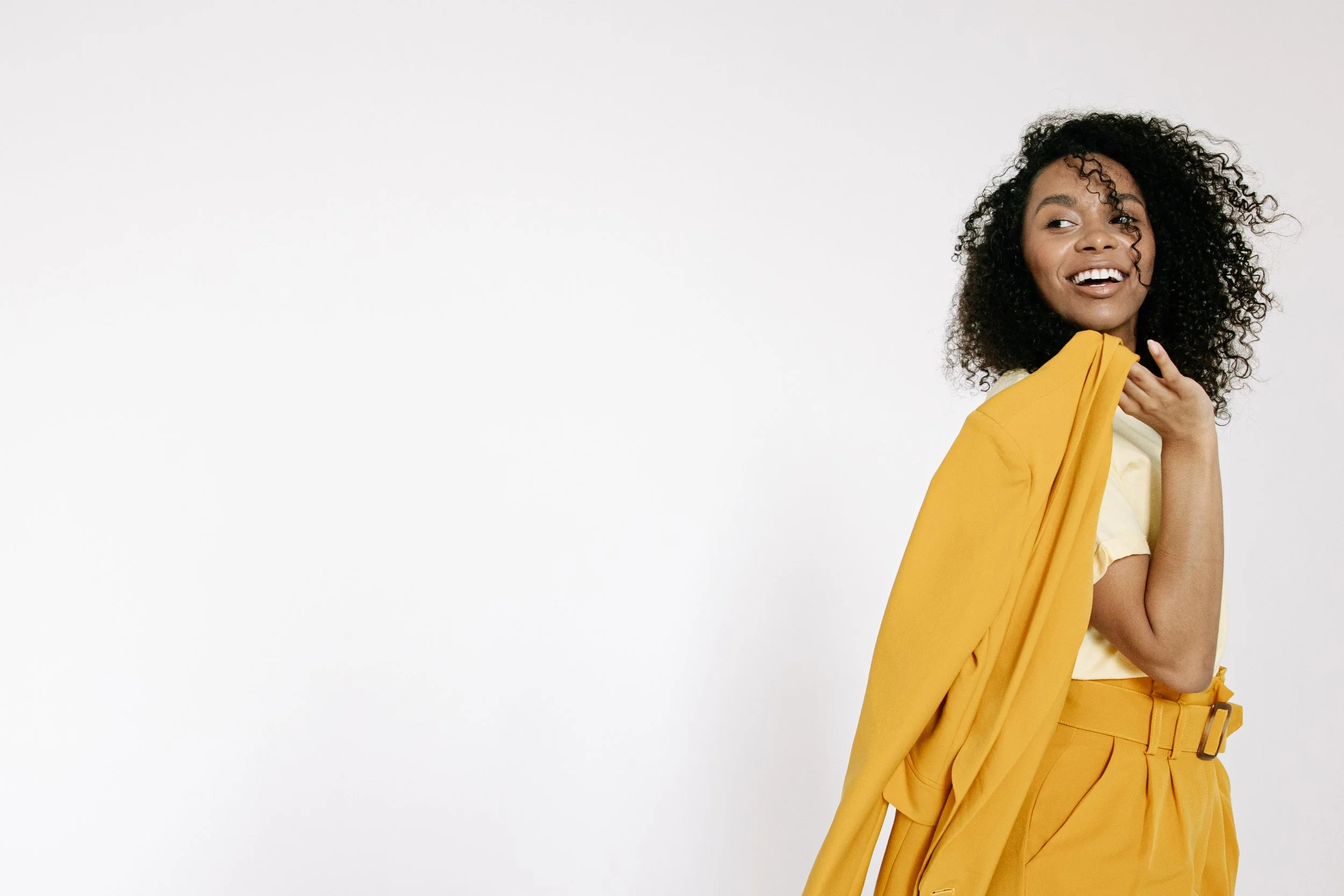 Young woman with curly hair smiling and wearing a yellow outfit against a plain white background.