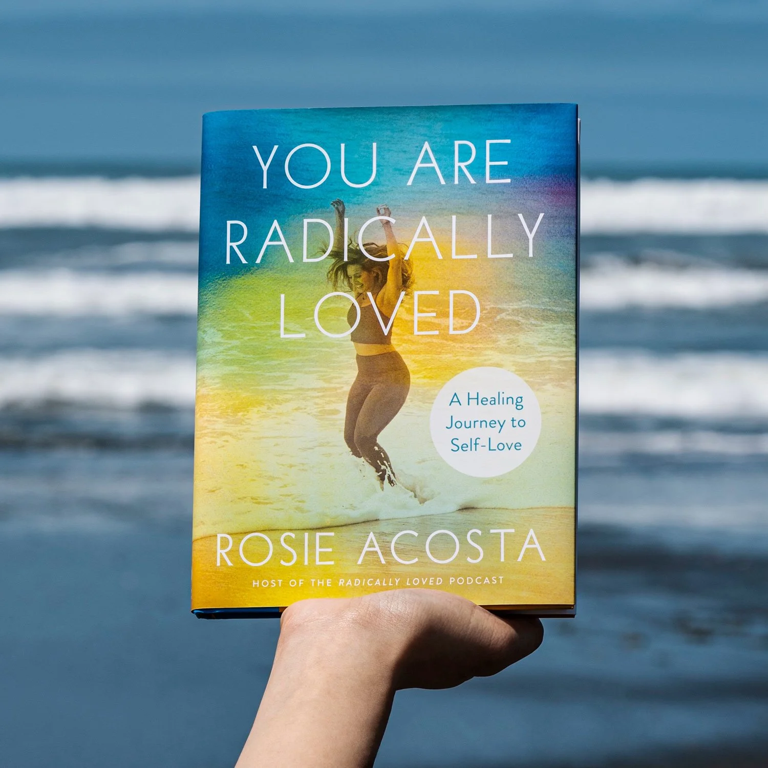 Book titled 'You Are Radically Loved' by Rosie Acosta held up against a beach background, with a woman jumping in the water on the cover.