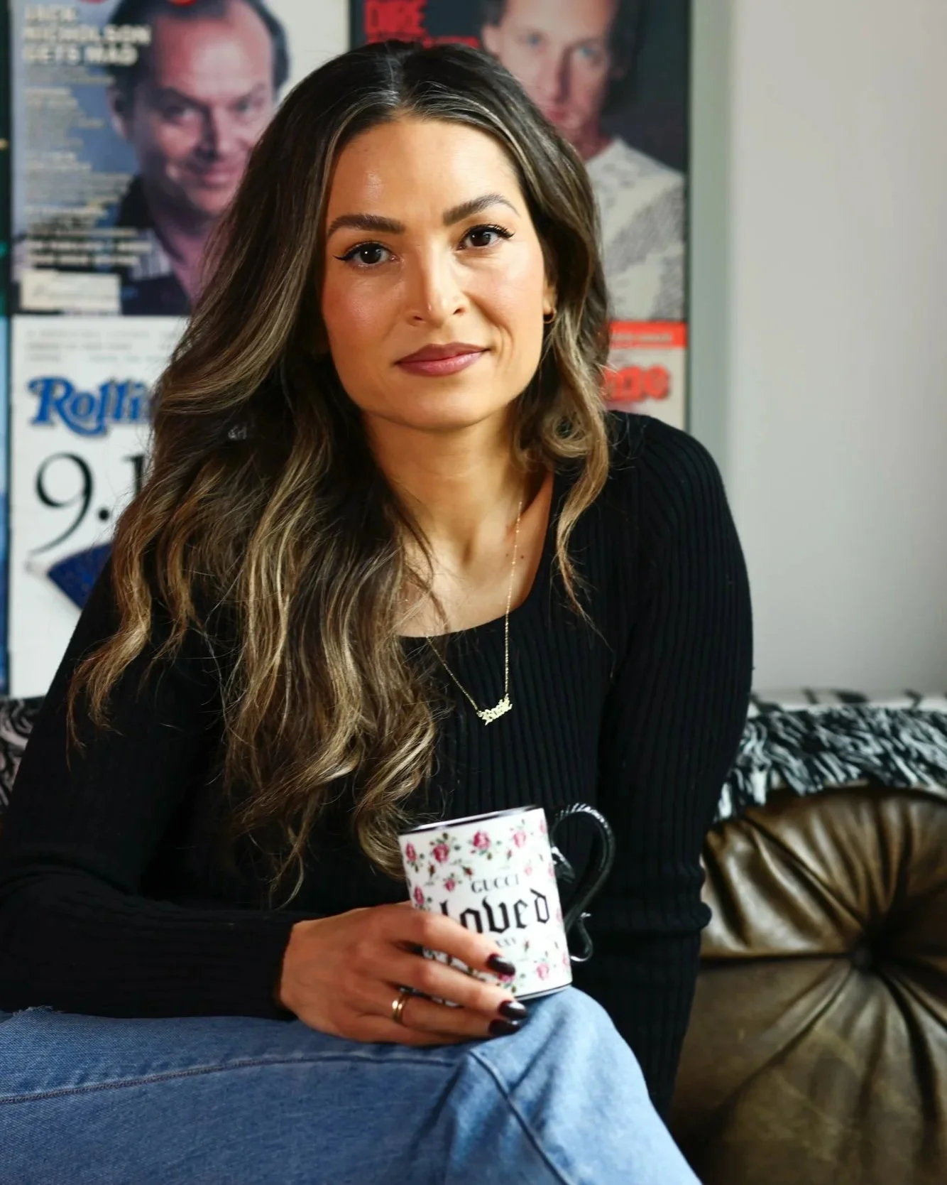 Rosie Acosta on a leather couch with a Radically Loved mug, in front of a wall of framed magazine covers